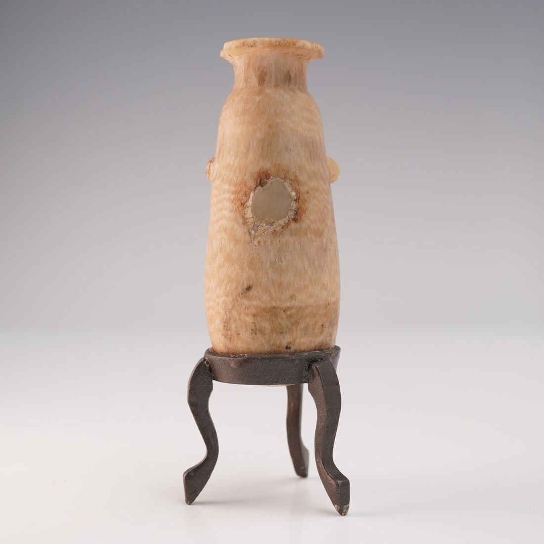 Ancient Alabaster Stone Flask with Metal Tripod Stand: A small stone flask featuring a cylindrical body with a narrow neck and flared rim, presented with a metal tripod stand. The surface displays a naturally variegated patina, with visible wear consisten
