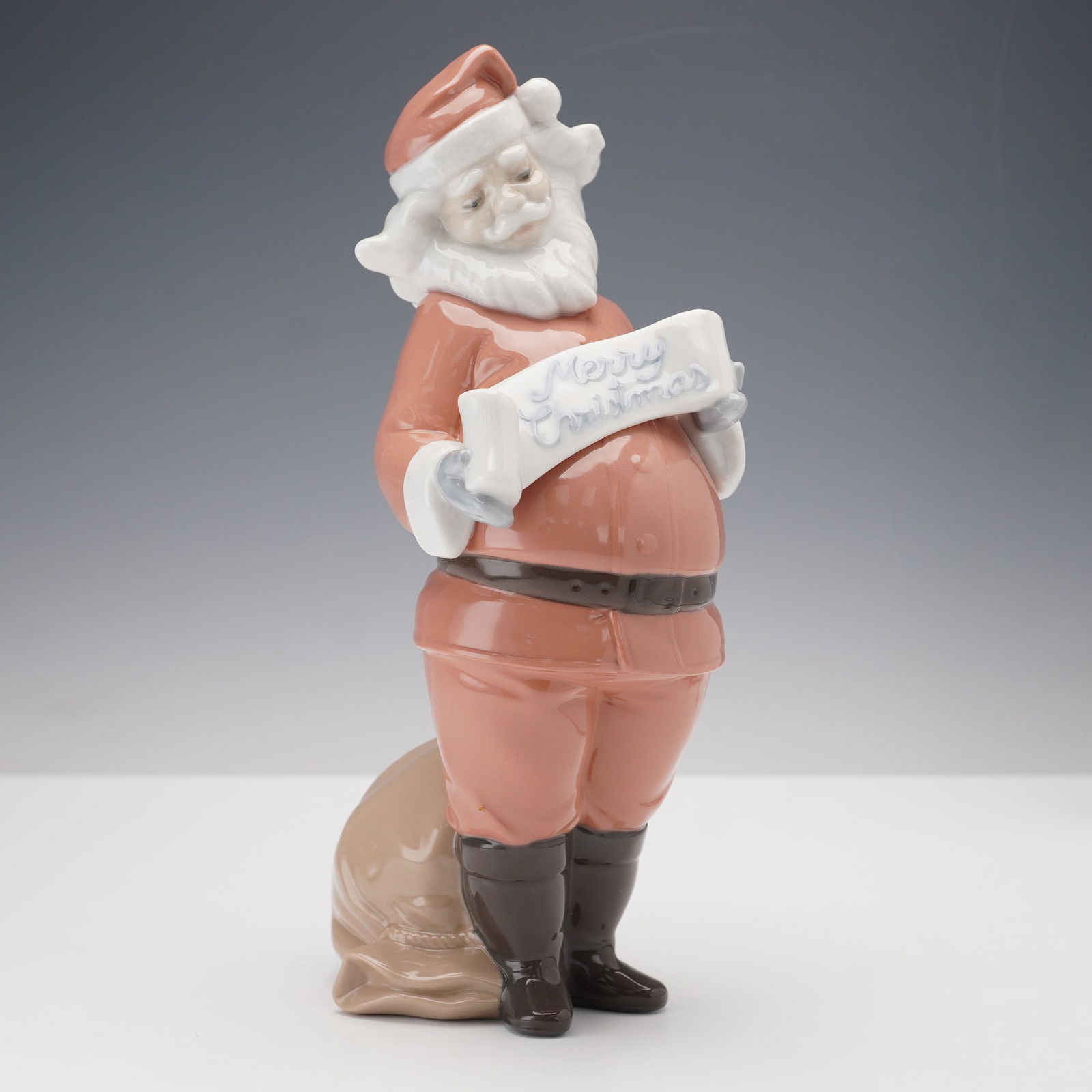 Nao by Lladro Figurine, Santa's Best Wishes 1399: This Nao by Lladro porcelain figurine, model number 1399, features Santa Claus holding a Merry Christmas banner. The piece is crafted with fine porcelain, showcasing soft pastel tones and delicate det
