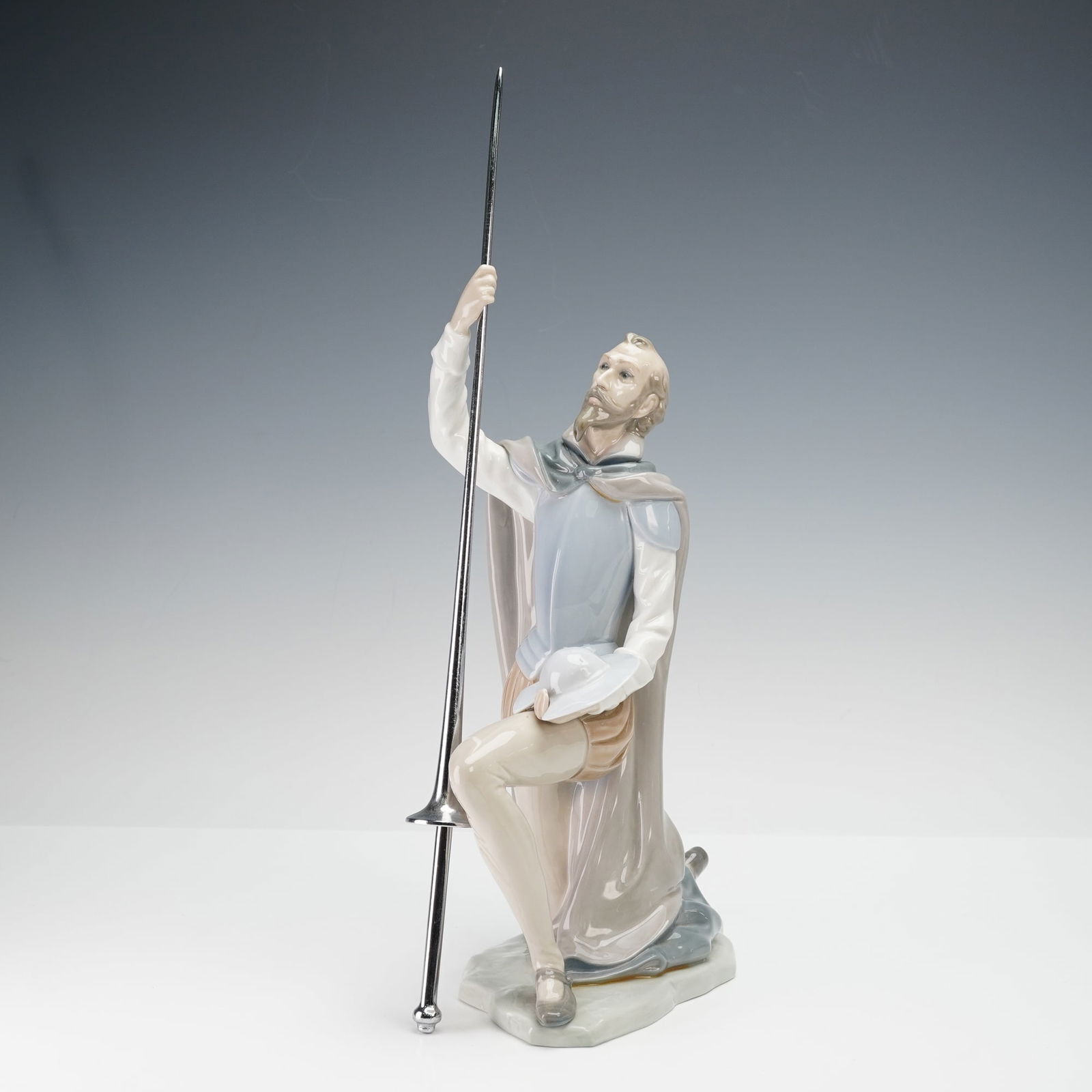 Lladro Porcelain Figurine, The Quest 1005224: Finely modeled Lladro porcelain figurine titled The Quest (1005224), depicting Don Quixote kneeling and gazing upward while holding a long lance and helmet. Rendered in soft pastel hues of gray, blue,