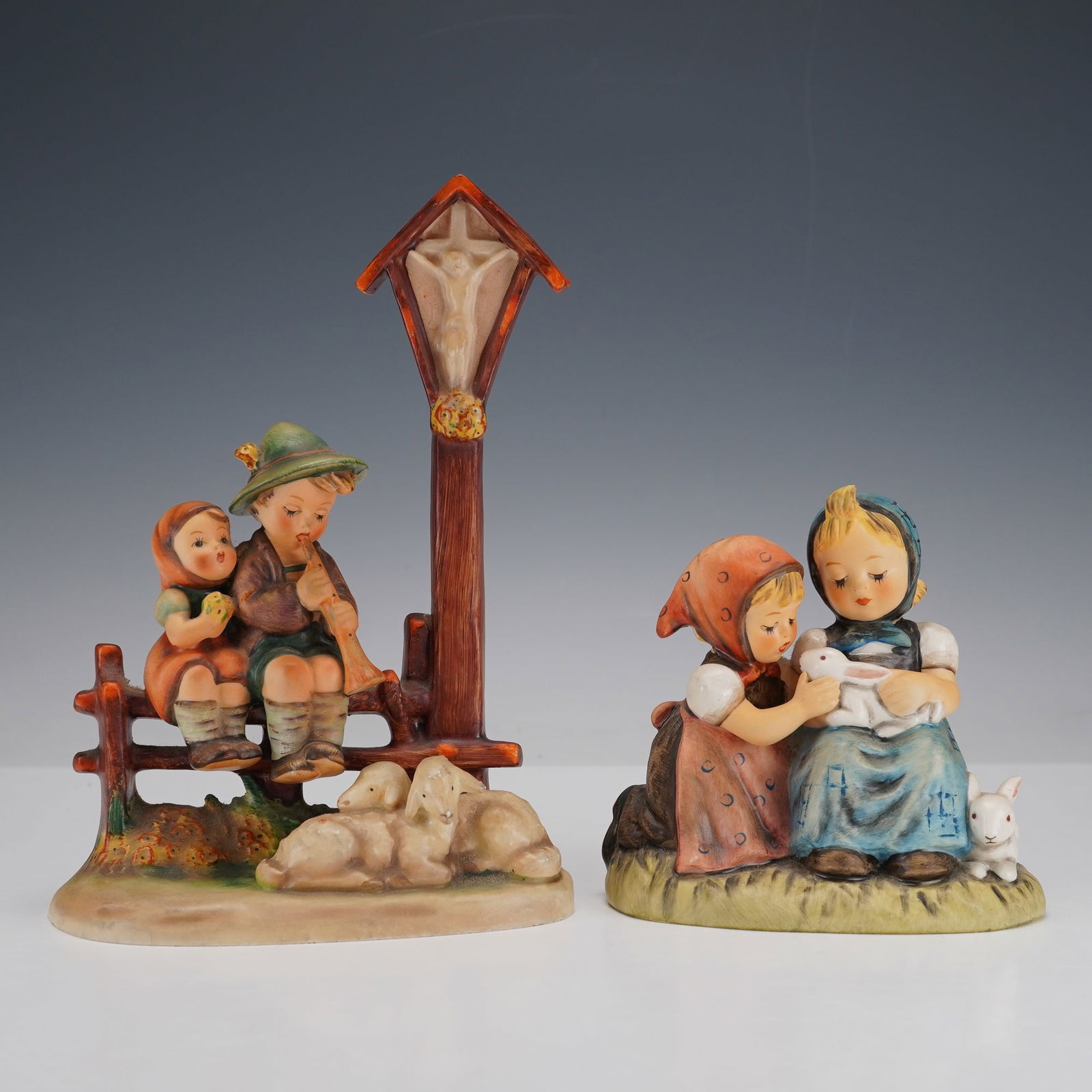 2pc Goebel Hummel Animal Companion Figurines: Finely hand-painted and showcasing the tender innocence of childhood. One depicts a boy and girl seated on a wooden fence beneath a crucifix, the boy playing a flute while sheep rest nearby. The secon