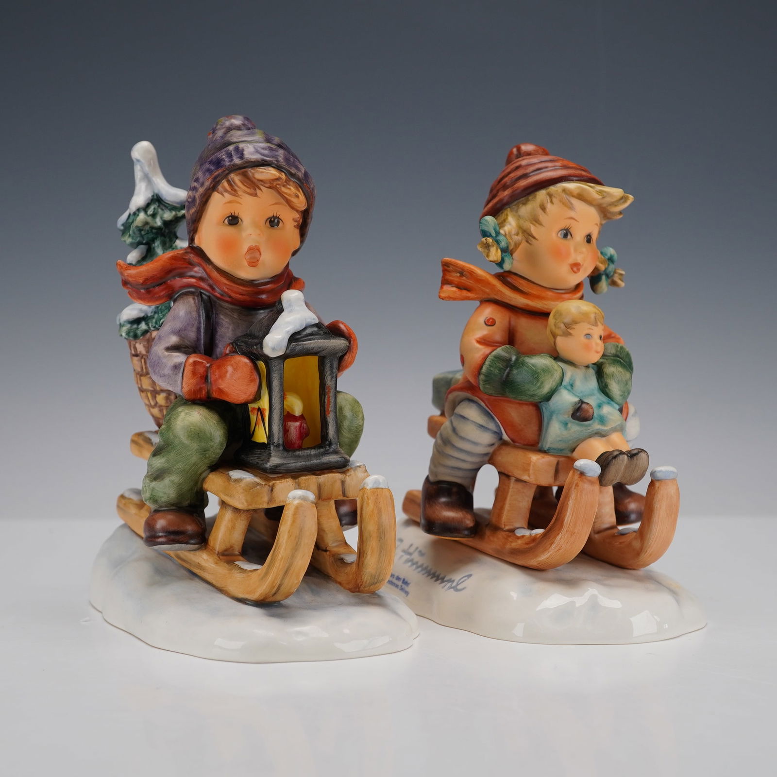 2pc Goebel Hummel Christmas Figurines: Two Goebel Hummel ceramic figures including Ride Into Christmas and Christmas Delivery. Depicts children sledding in snowy scenes, one carries a lantern and fir tree on a toboggan and the other child