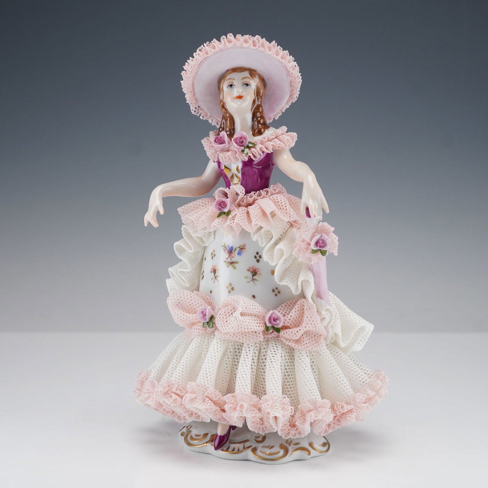 Dresden Porcelain Lady in Pink Gown Lace Figurine: A finely detailed Dresden porcelain lace figurine depicting a lady in a tiered pink and white gown with floral accents and a wide-brimmed hat. Her ruffled dress features delicate applied porcelain lac