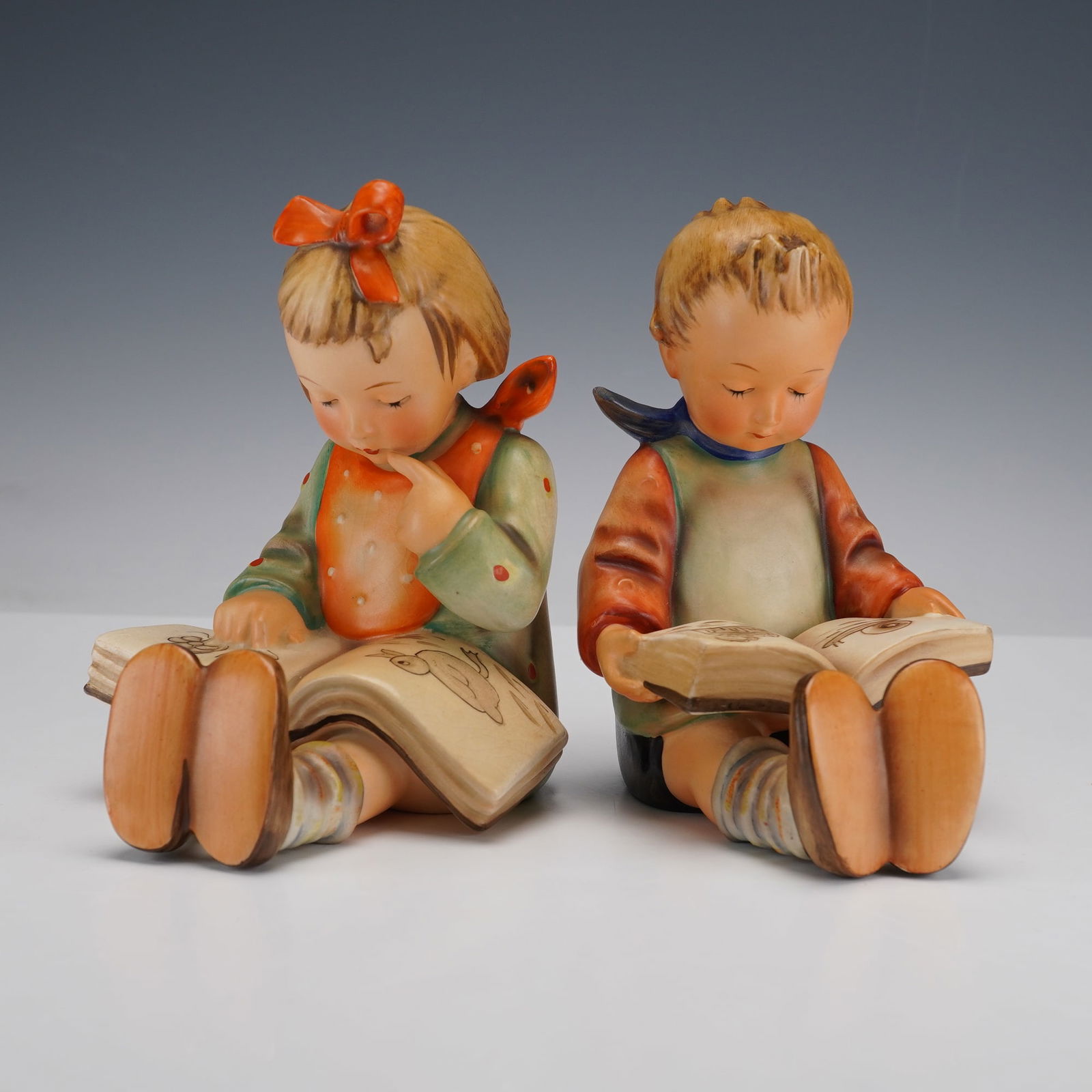 Pair of Goebel Hummel Bookends, Bookworm Boy and Bookworm Girl: Pair of bookends modeled as a seated girl and boy absorbed in illustrated books. Each depicted in hand painted attire. M. I. Hummel stamped to the back of the bookends. Goebel backstamp. Dimensions: 4