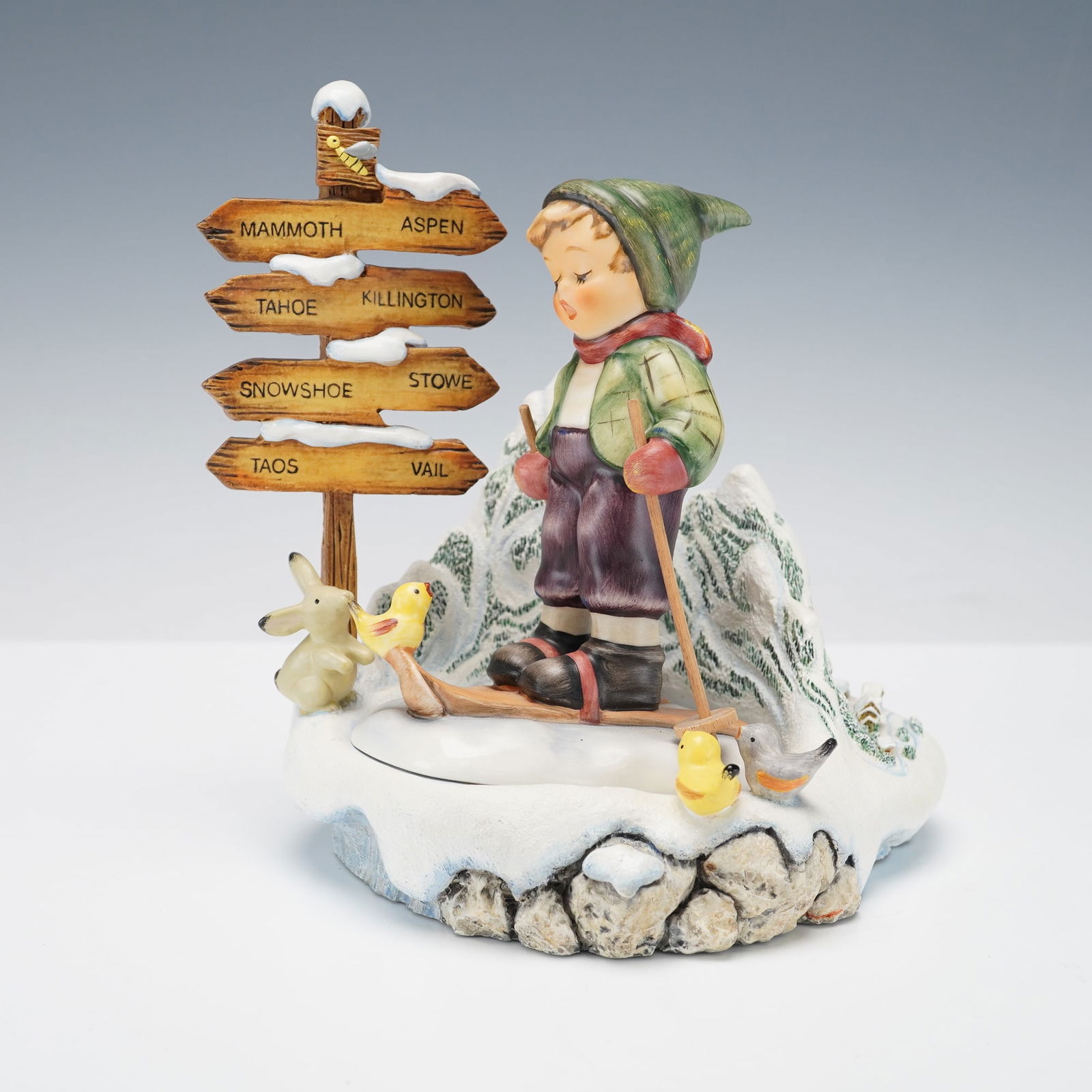 2pc Goebel Hummel Figurine & Hummelscape, Winter Adventure & Slalom Slopes: This Goebel Hummel display pairs the figurine Winter Adventure with the HummelScape base Slalom Slopes. The boy stands on skis with a small bird perched near his feet, dressed warmly with a green cap