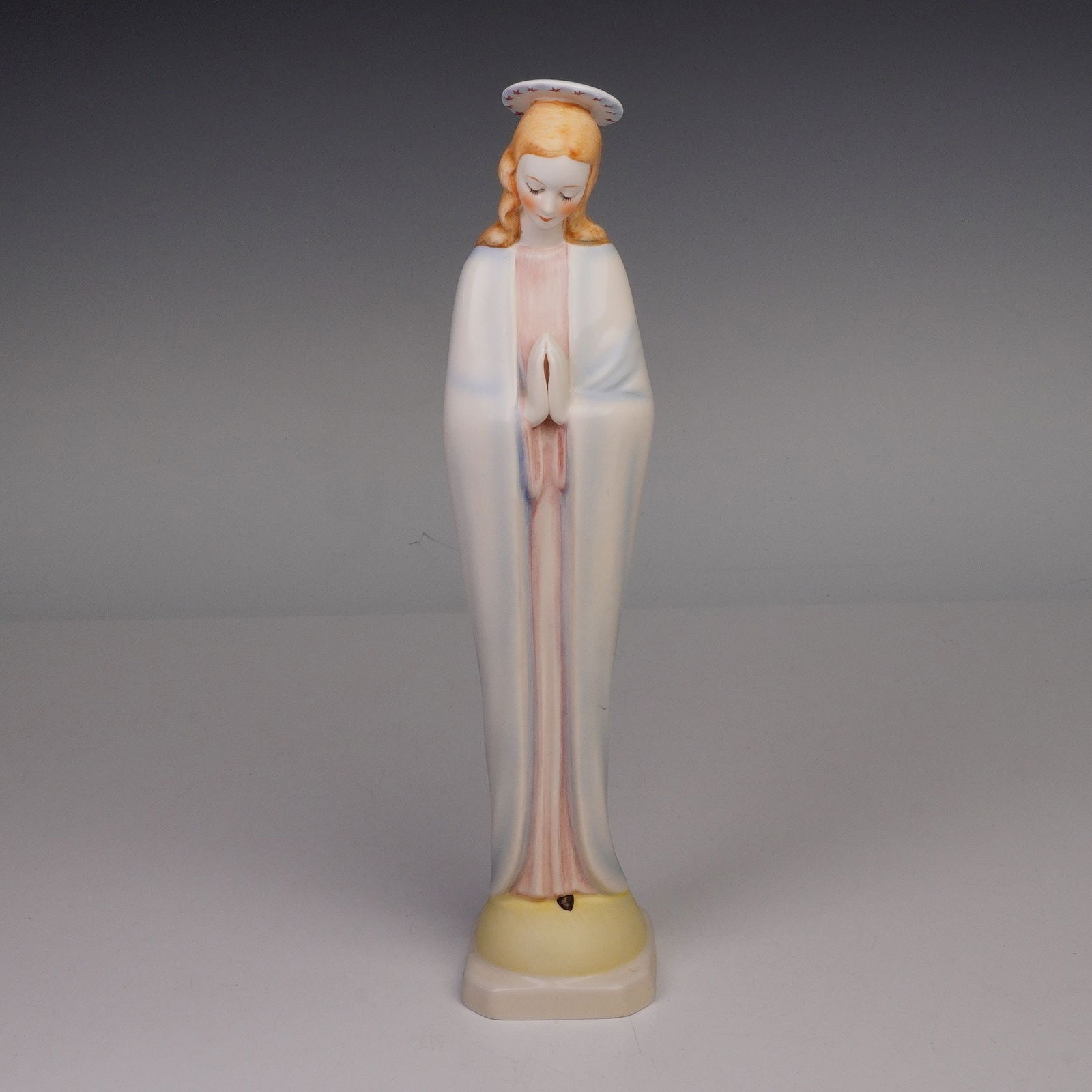 Goebel Hummel Figurine, Madonna: A Goebel Hummel porcelain figurine depicting the Madonna, hands clasped in prayer, with soft pastel tones of blue, pink, and cream. The figure stands on a shaped base and retains its original halo. Ma