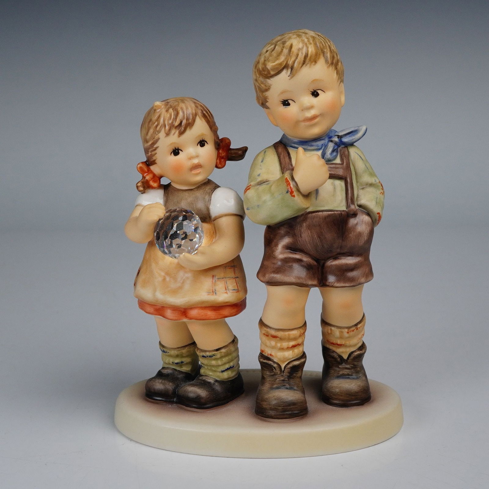Goebel Hummel Figurine, She Caught It!: Depicting a young boy and girl standing side by side, with the girl holding a faceted crystal ball in her hands. Hand-painted in soft pastel tones, the piece exemplifies the charming innocence charact