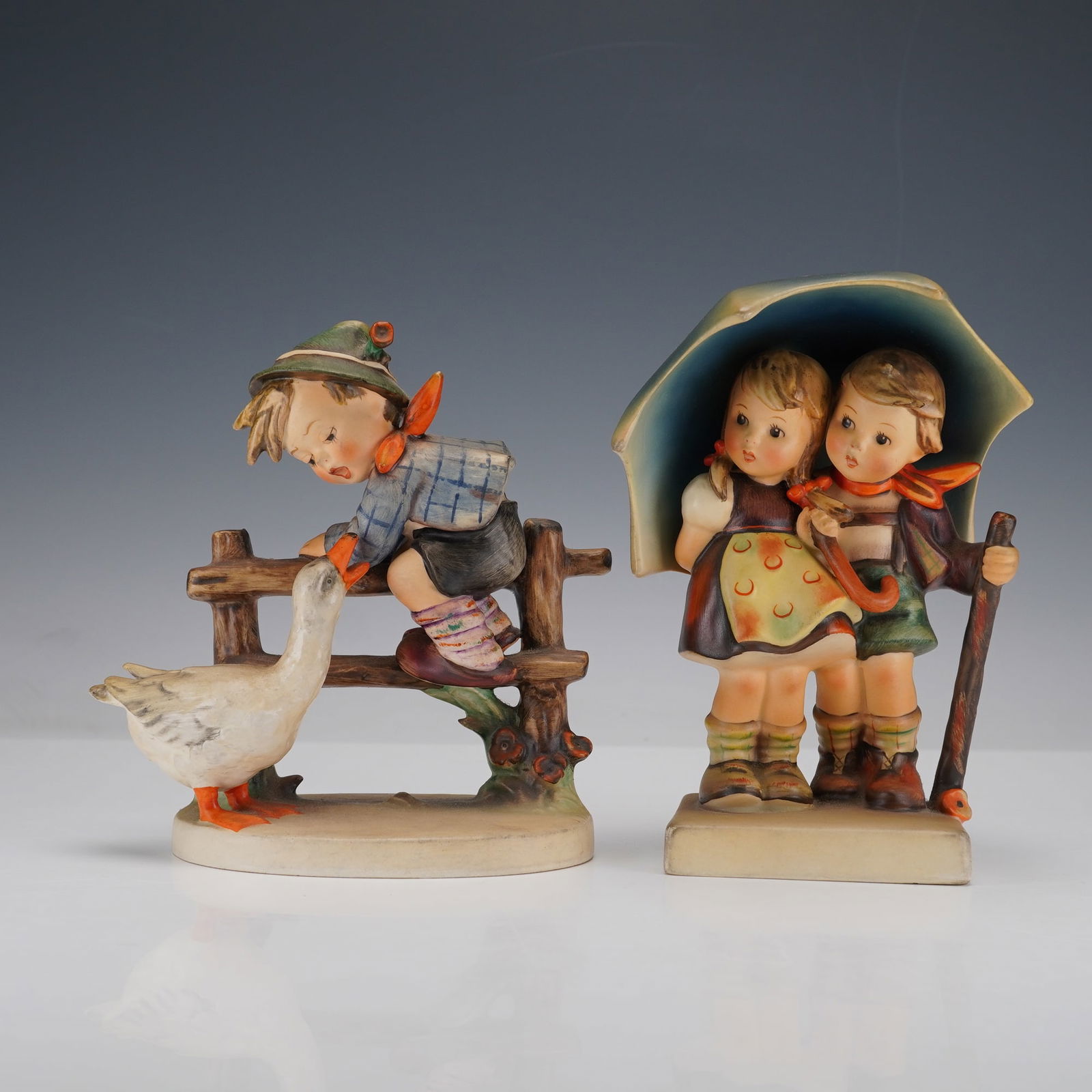 2pc Goebel Hummel Figurines, Barnyard Hero and Stormy Weather: A charming pair of Hummel figurines including Barnyard Hero, showing a young boy perched on a fence as a curious goose approaches, and Stormy Weather, featuring a boy and girl huddled together beneath