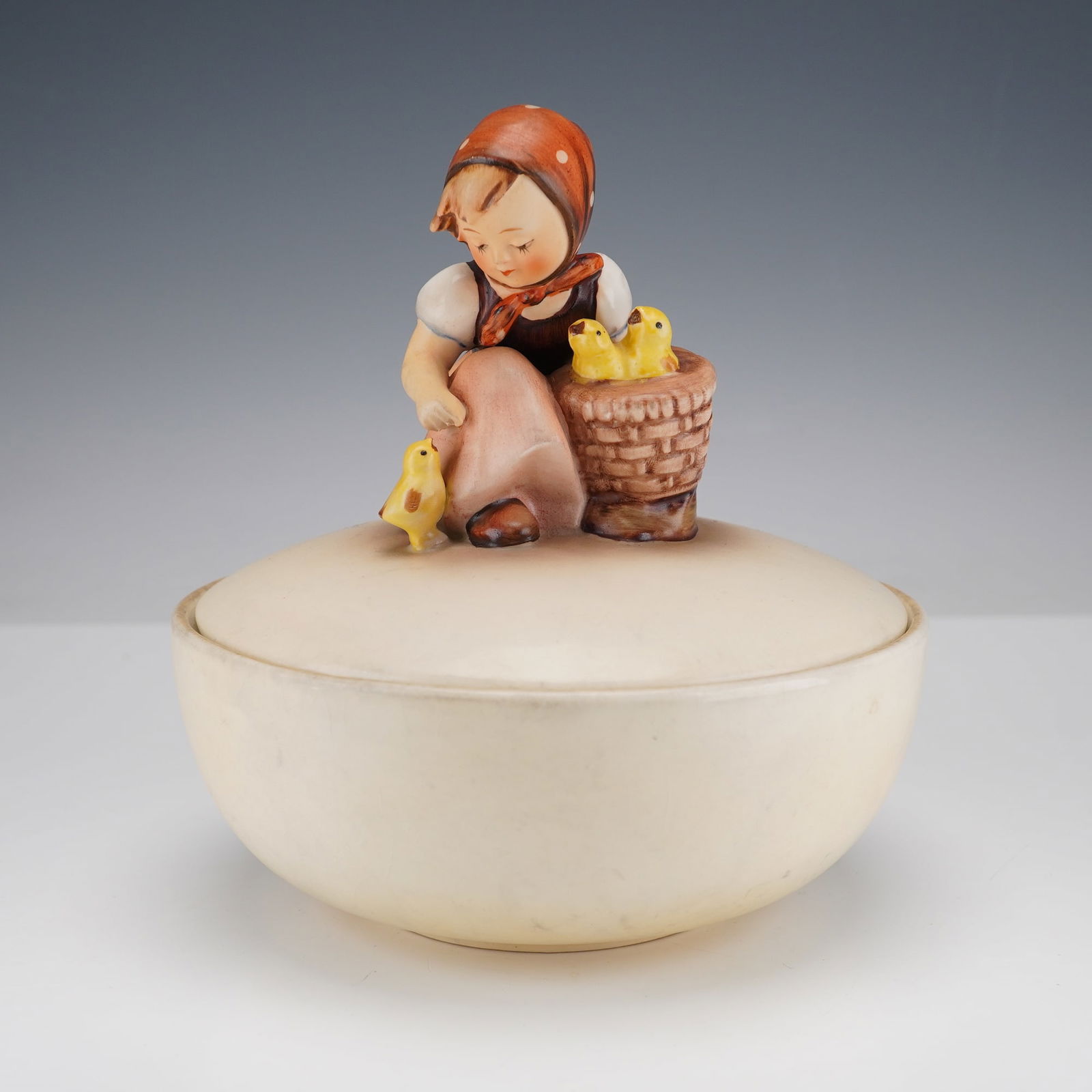 Goebel Hummel Coverd Candy Dish, Chick Girl: A delicately hand-painted young girl seated beside a basket of yellow chicks, with one chick perched near her hand. The figure is mounted atop a smooth, rounded lidded bowl with a creamy ivory glaze,