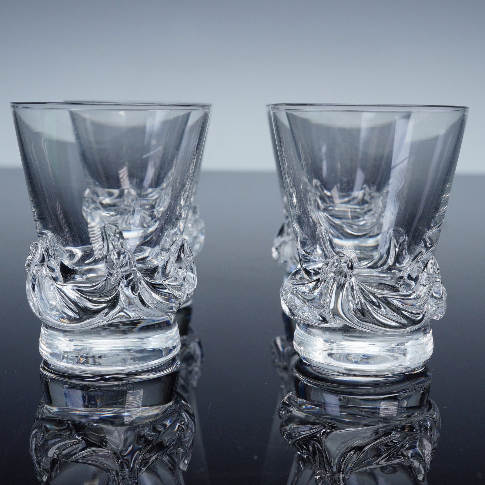 4pc Daum Sorcy Flat Tumbler Glasses: These finely crafted crystal glasses feature a striking sculptural base with swirling, organic forms that catch and reflect the light beautifully. The sleek, tapered bowls contrast elegantly with the