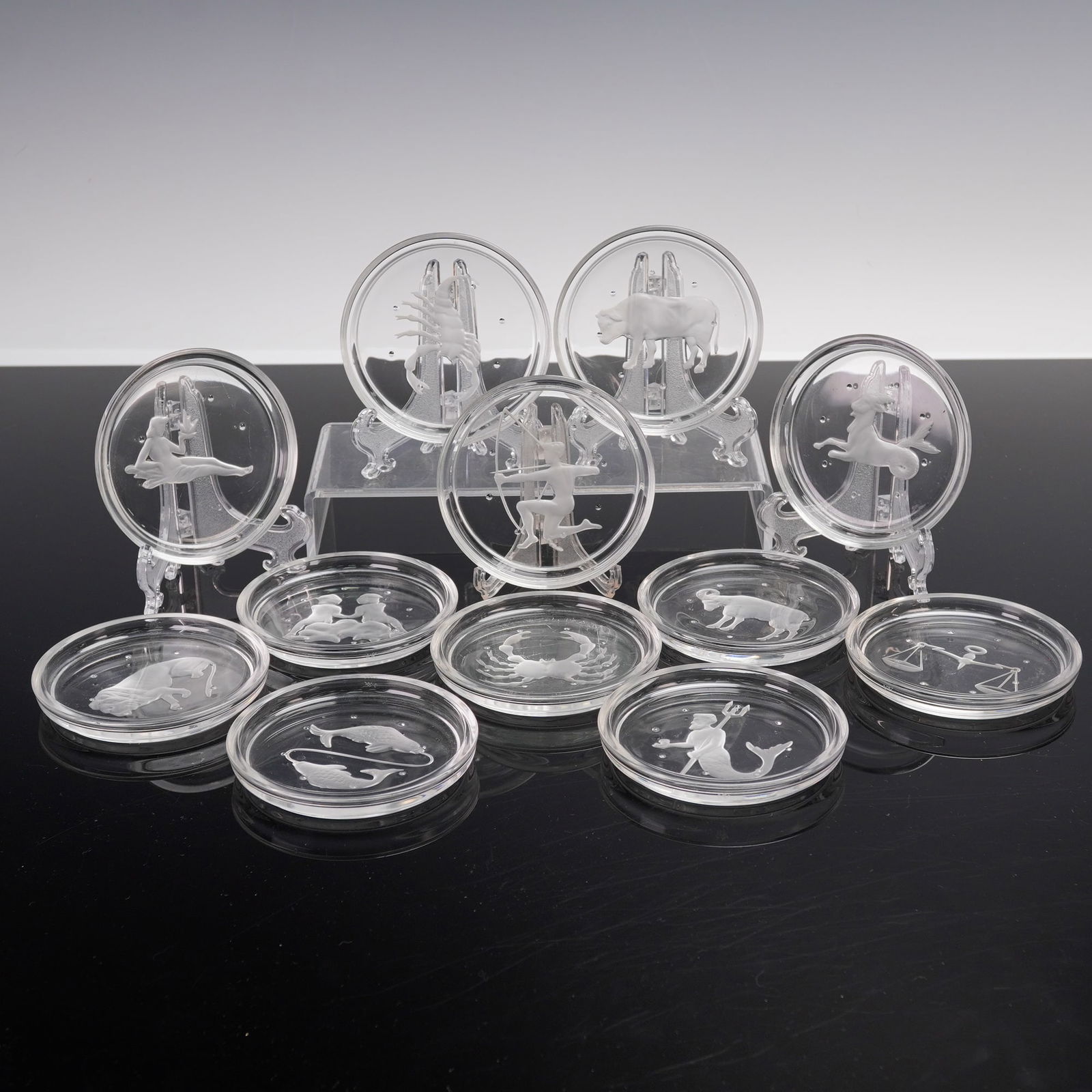 12pc Val Saint Lambert Zodiac Glass Coasters: A set of twelve round glass coasters by Val Saint Lambert, each finely decorated with an intaglio design representing a sign of the Zodiac. Signed pieces from this renowned Belgian glassworks, combini