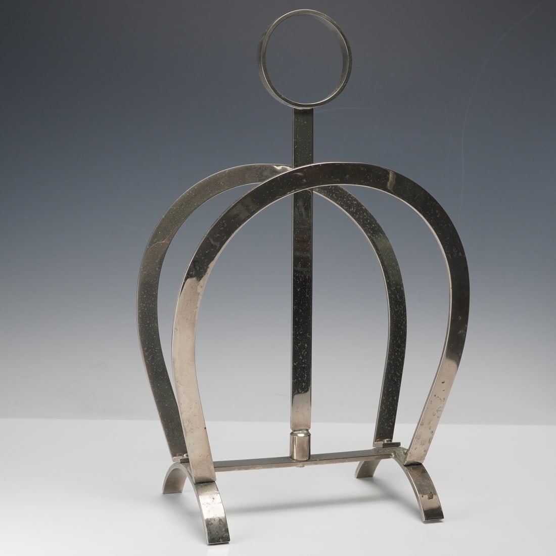 Modernist Metal Magazine Rack: A striking modernist magazine rack crafted from polished metal, featuring an arched horseshoe-shaped frame and circular finial handle. Its clean geometric form and balanced proportions highlight a ref