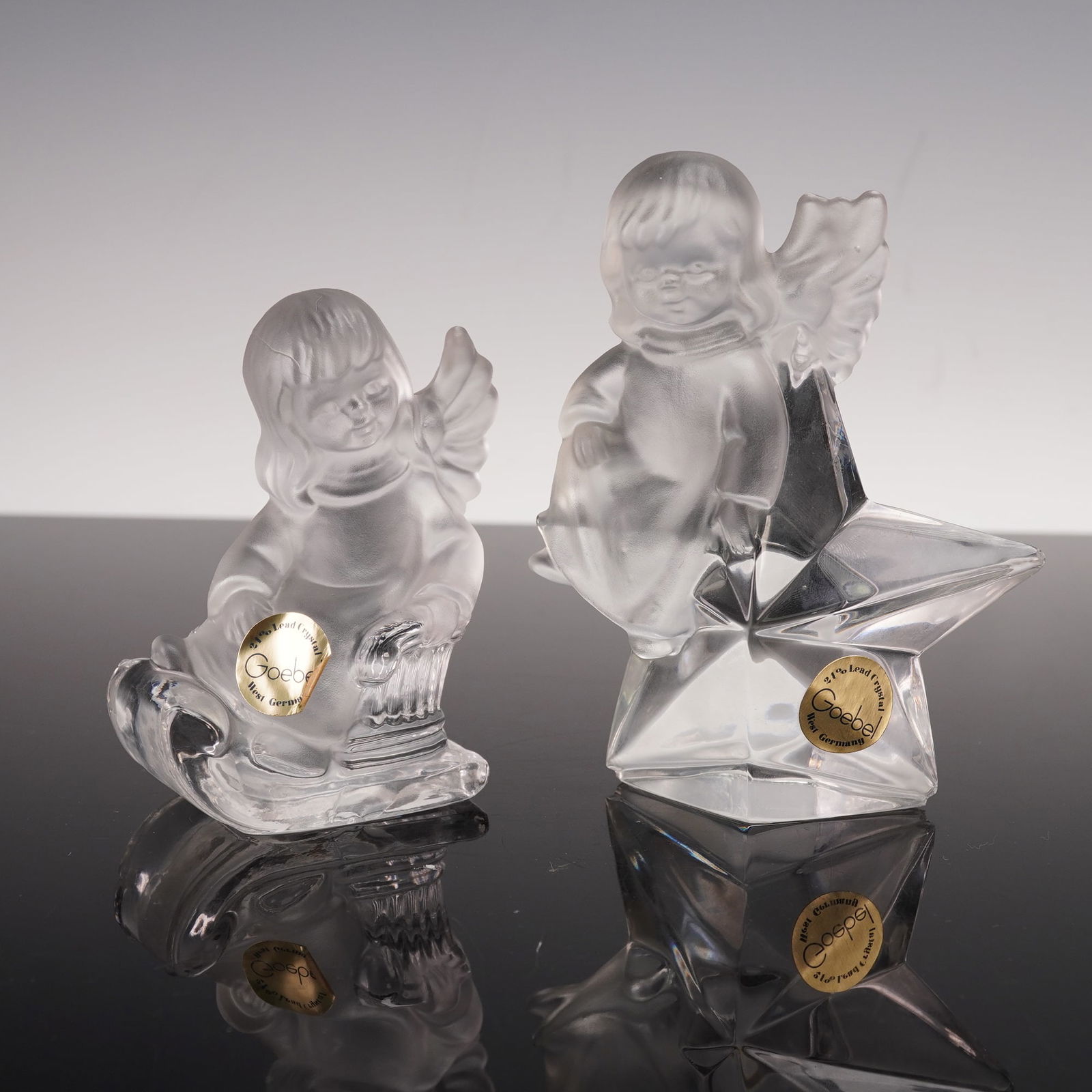 2pc Goebel Hummel Crystal Angel Figurines: These two Goebel crystal angel figurines are made of frosted glass with clear accents that reflect light beautifully. One angel rests upon a faceted star while the other sits on a curved sled. Both fi