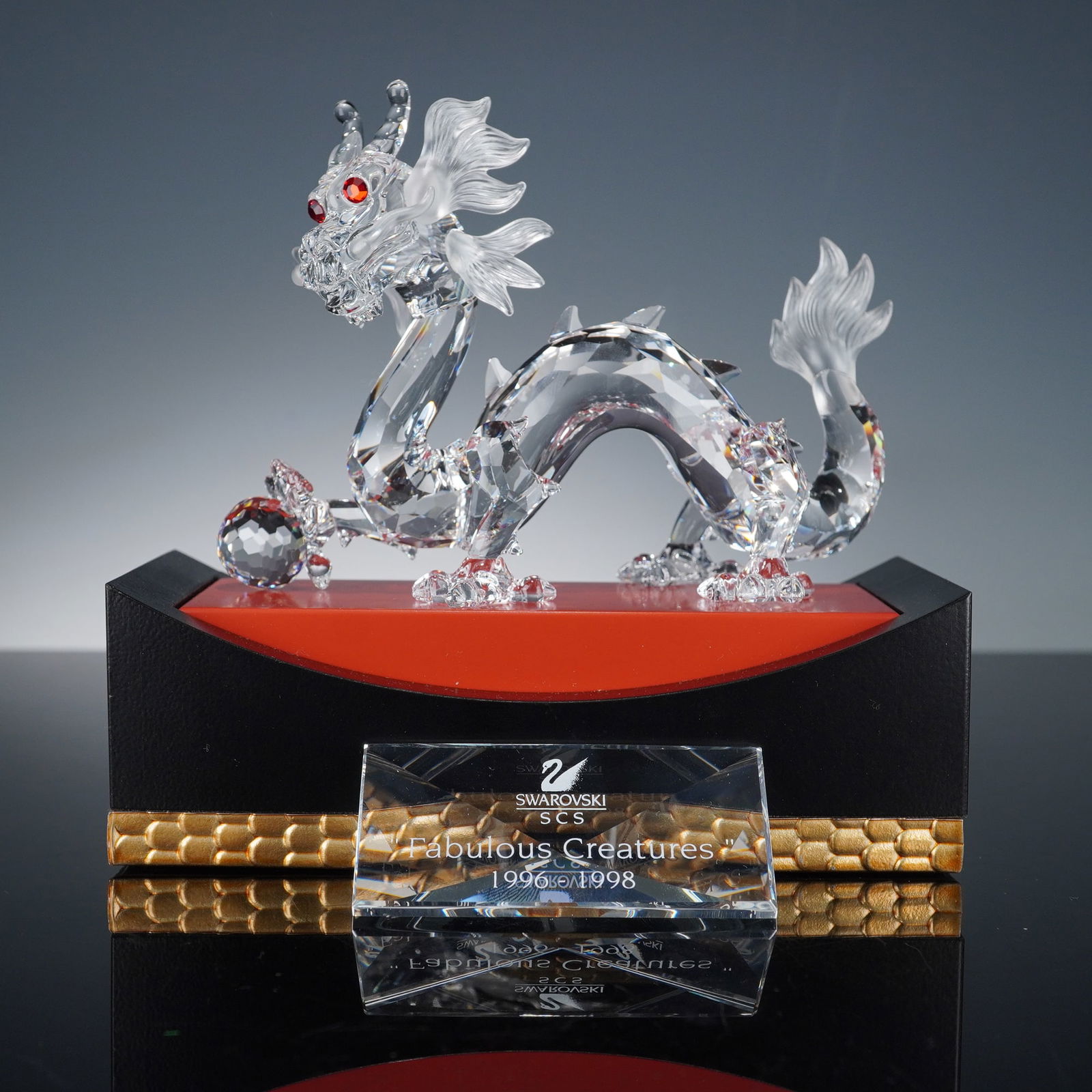 2pc Swarovski Fabulous Creatures Crystal Figurine, Dragon & Plaque: Depicting a gracefully arched dragon crafted in faceted clear crystal with frosted detailing and red crystal eyes, poised beside a crystal sphere. Accompanied by a coordinating crystal plaque etched w