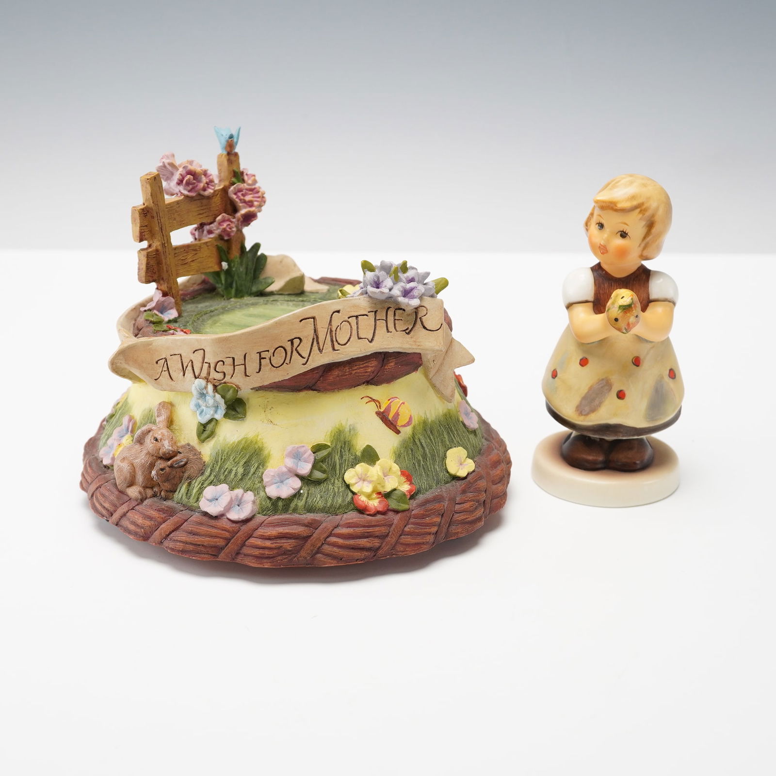 2pc Goebel Hummel Music Box and Figurine, A Wish for Mother: Two-piece Goebel Hummel set including A Wish for Mother music box base adorned with flowers, a fence, and banner plus a porcelain figurine of a young girl holding flowers. The music box plays I Just C