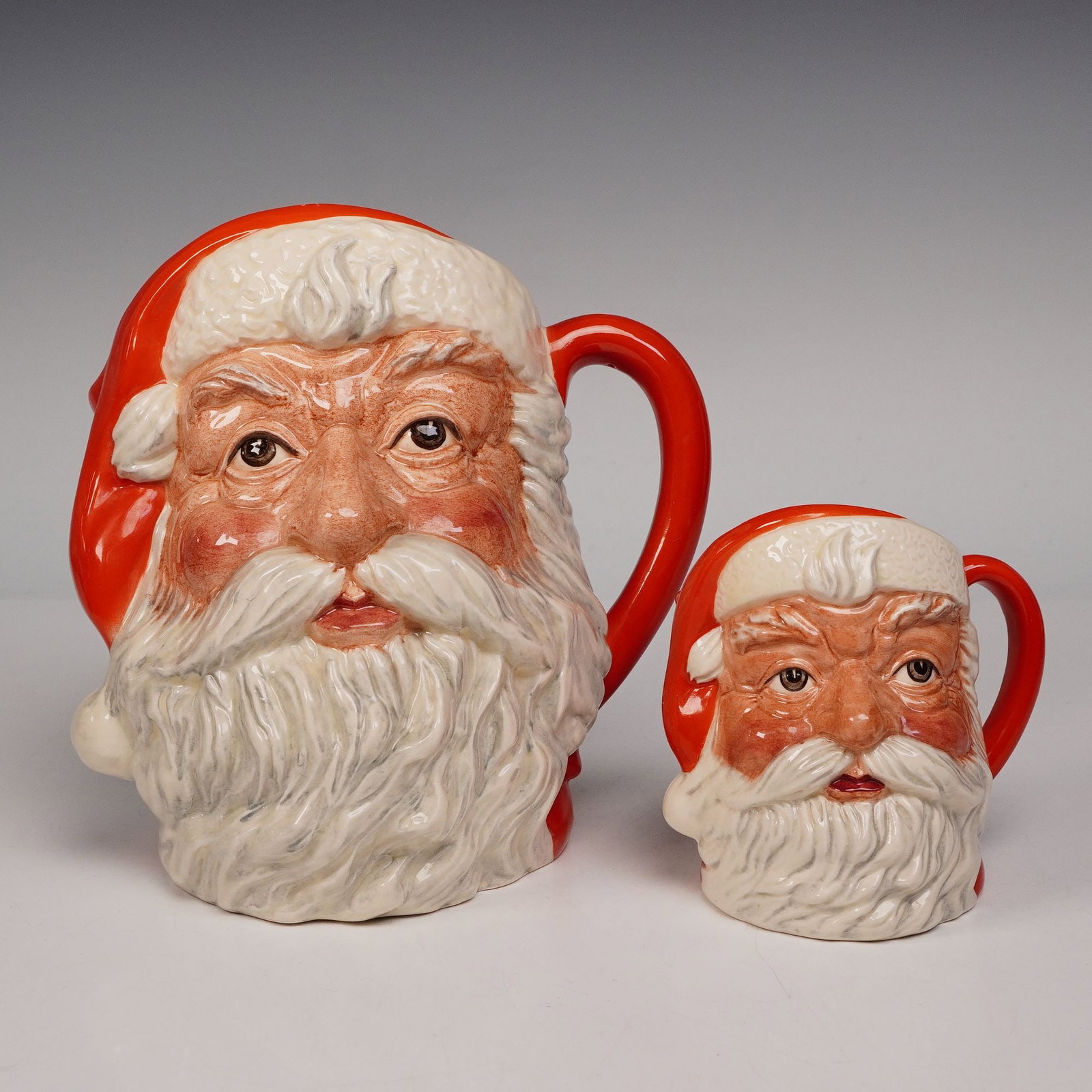 2pc Royal Doulton Character Jugs, Large and Small Santa Claus: A festive collection of two character jugs, comprising one large D6704 and one small version D6705. Each jug portrays the traditional visage of Santa Claus, with a flowing white beard, red cap, and ex