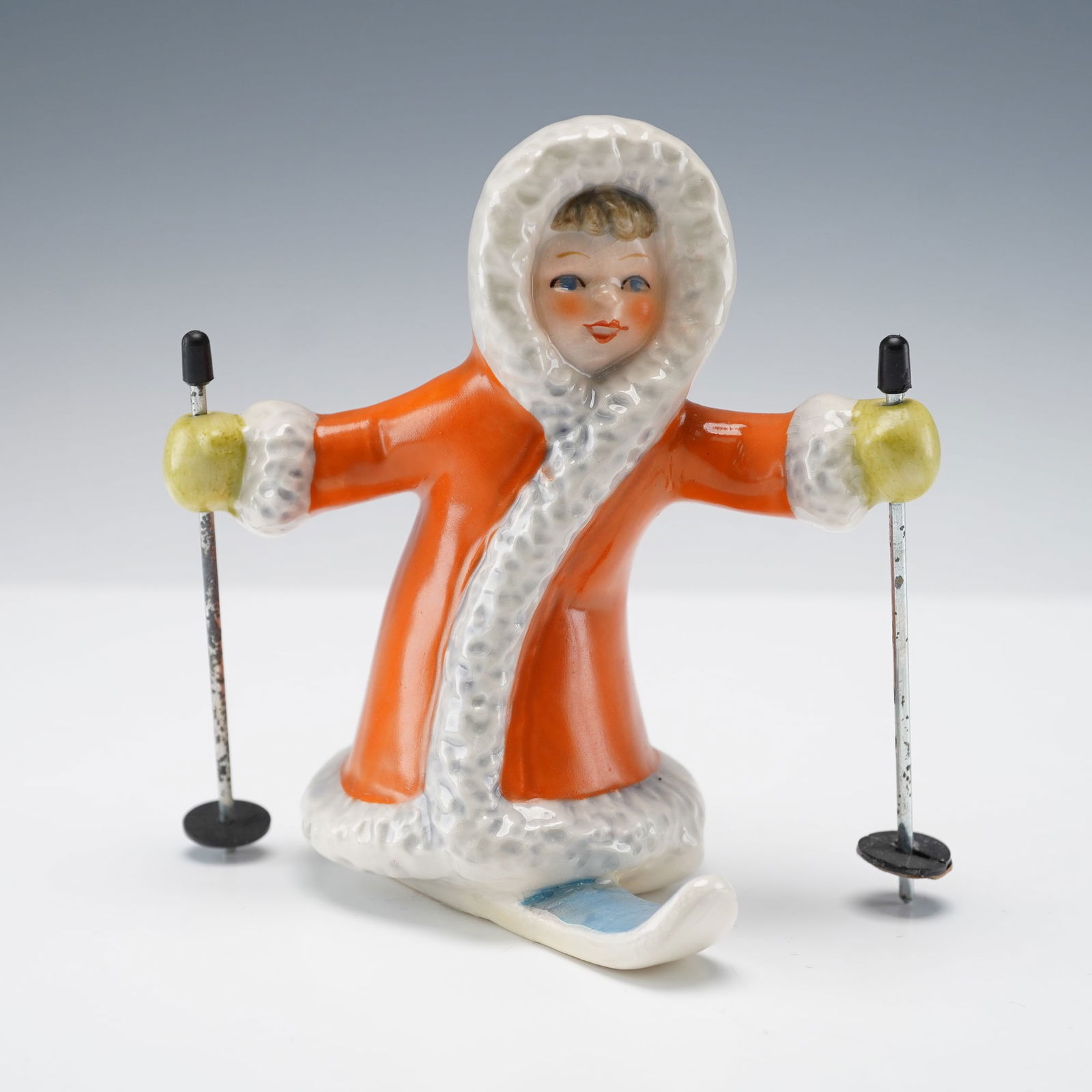 Goebel Hummel Figurine, Skiing Girl: This charming Goebel Hummel porcelain figurine shows a cheerful girl gliding on skis, dressed warmly for a winter day. She wears a bright orange coat with soft white trim and yellow mittens and holds