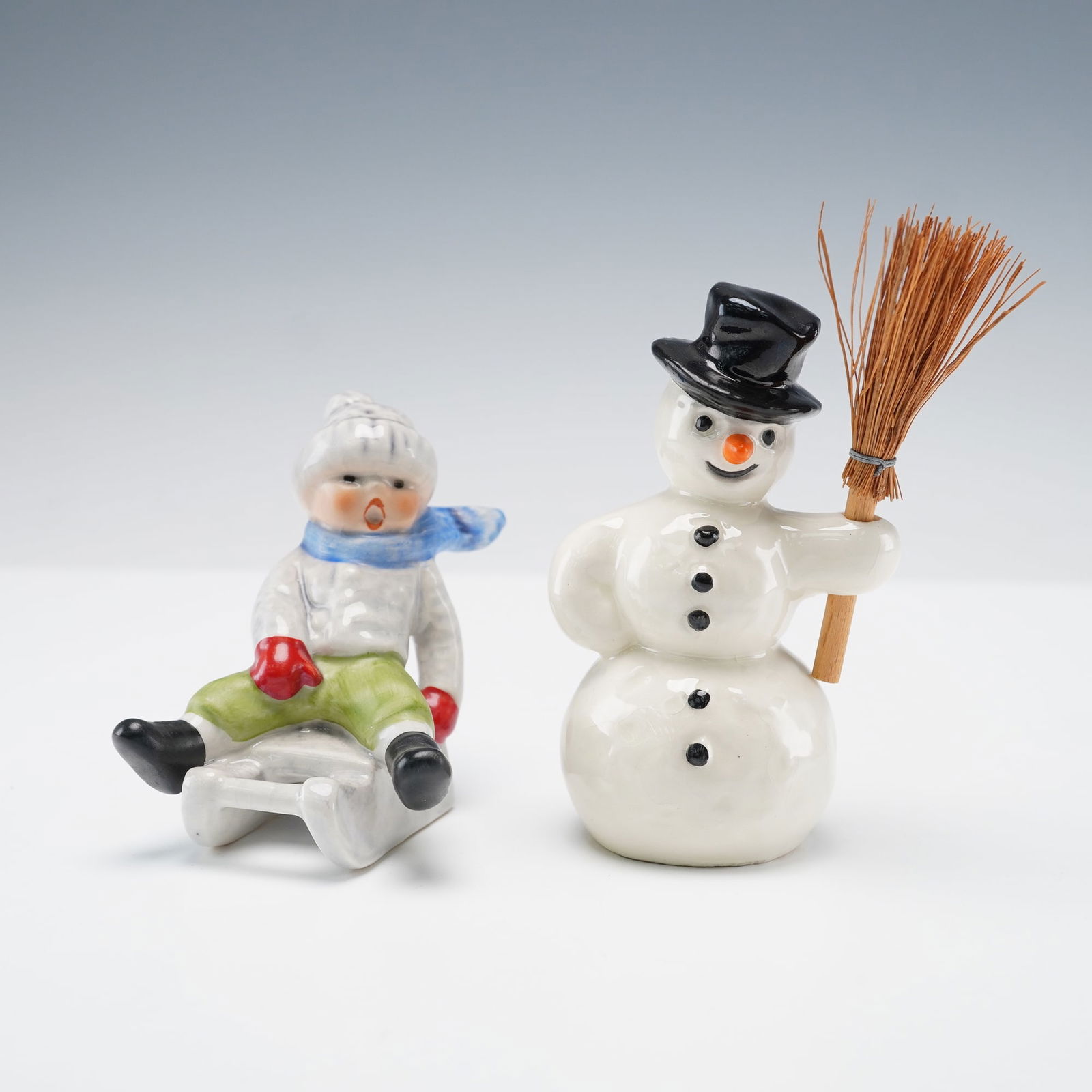 2pc Goebel Hummel Figurines, Snowman and Boy on Sled: This delightful pair of Hummel figures captures a cheerful winter scene. The snowman stands with a friendly smile, wearing a classic top hat and holding a broom, his round shape and simple details evo