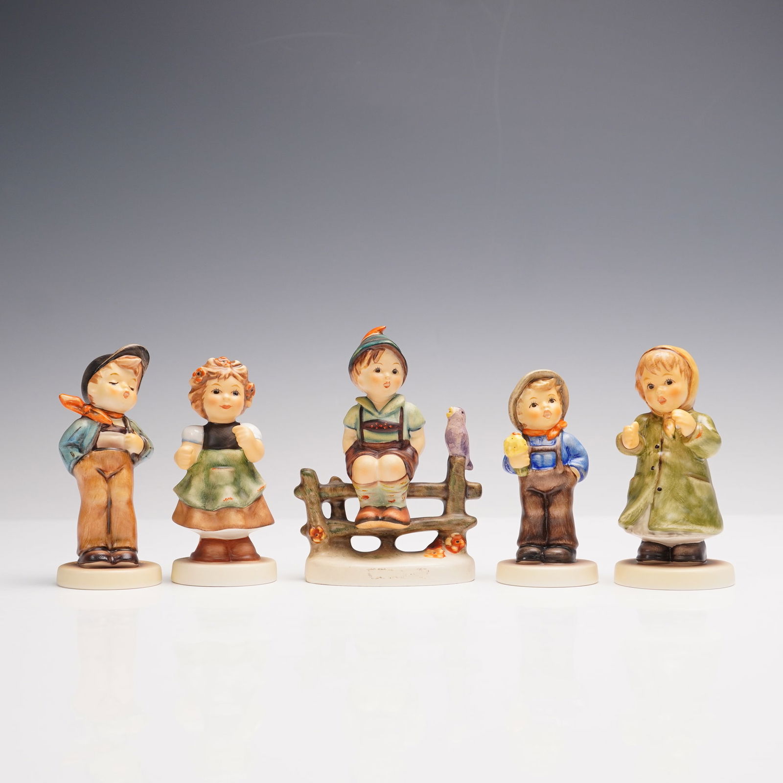 5pc Goebel Hummel Figurines, Whimsical Village Children: A set of five Goebel Hummel porcelain figurines, all hallmarked. The group features a boy with his hand in his pocket and a scarf at his neck, a young girl in a dress and apron with flowers in her hai