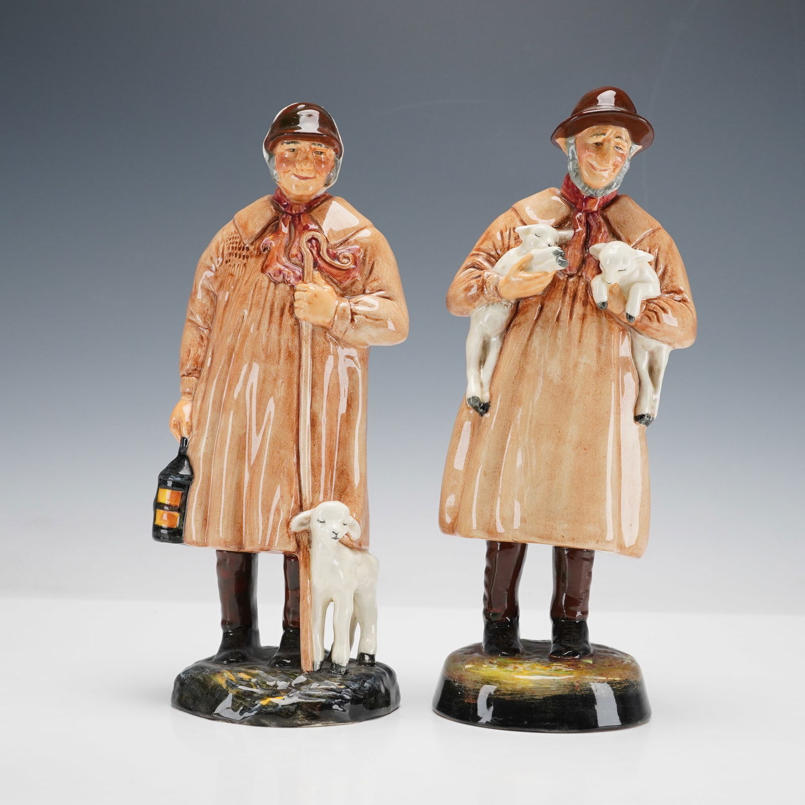 2pc Royal Doulton Figurines, The Shepherd HN1975 and Lambing Time HN1890: Two figurines titled The Shepherd HN1975 and Lambing Time HN1890. Each depicts a rustic country shepherd dressed in a traditional smock, one holding a lantern beside a resting lamb, the other cradling