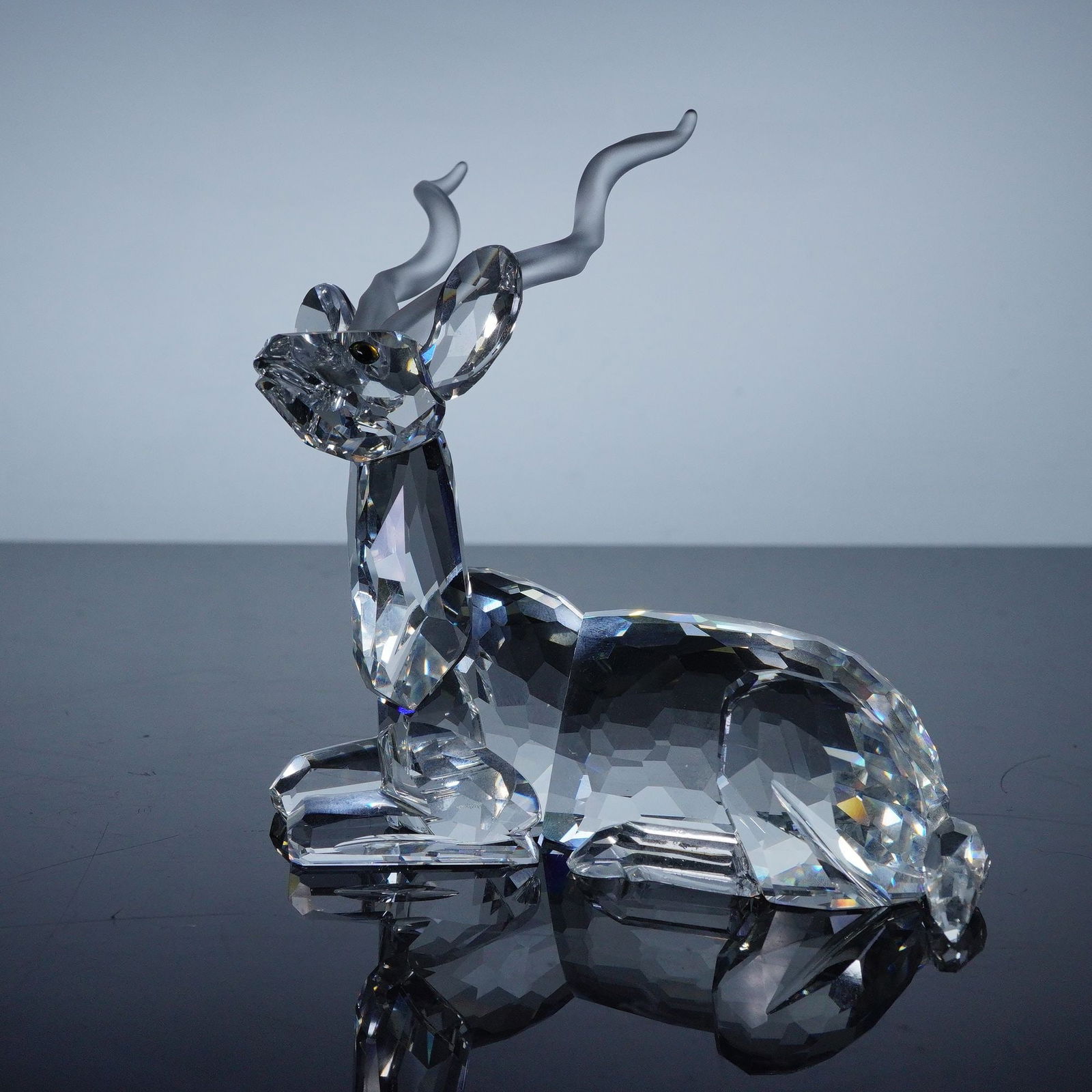 Swarovski Inspiration Africa Crystal Figurine, Annual Edition Kudu: A masterfully crafted clear crystal figurine depicting a reclining kudu, showcasing Swarovski's renowned precision-cut facets that capture and refract light brilliantly. The piece features gracefully