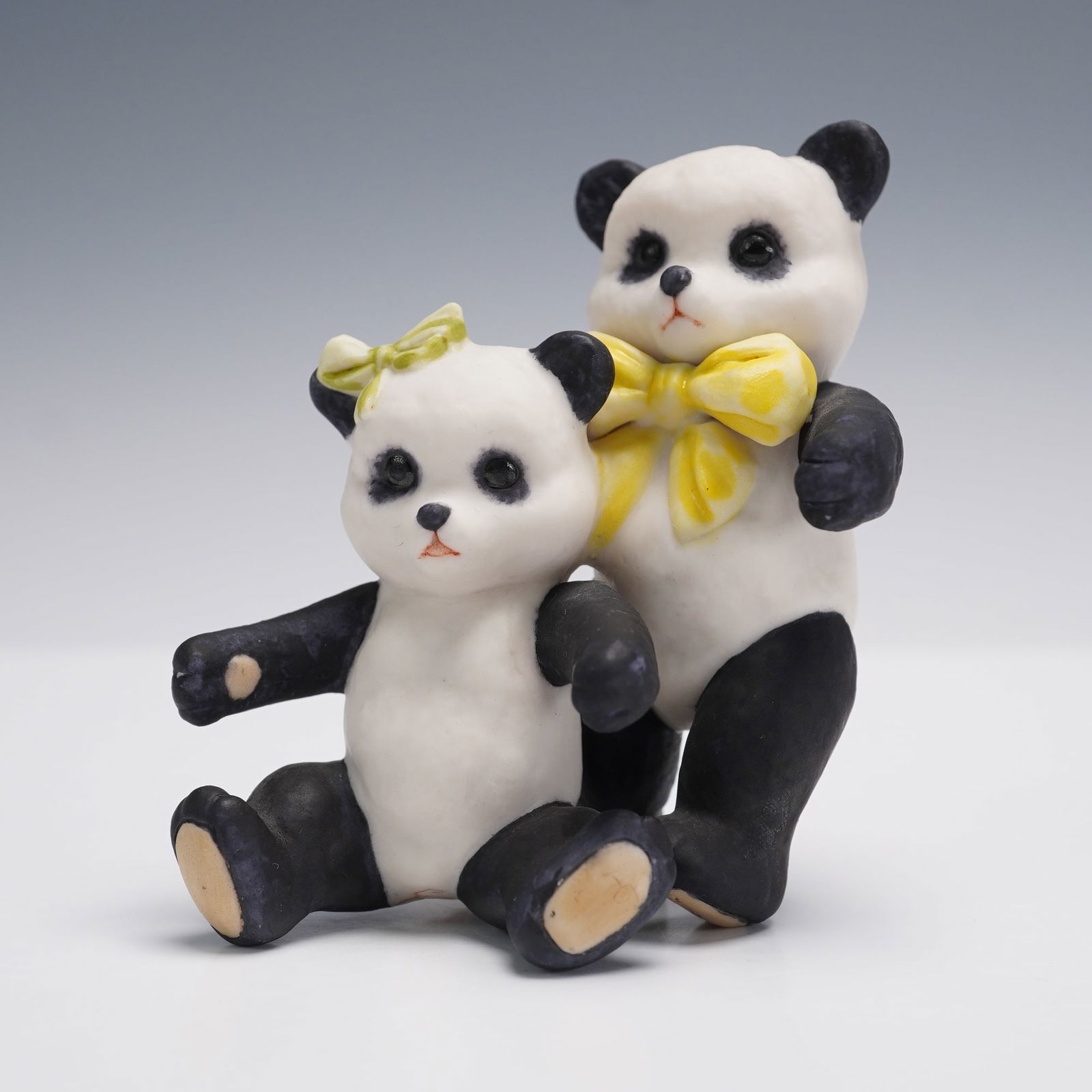 Cybis Porcelain Panda Pair Figurine: Cybis porcelain figurine depicting two charming panda bears, one seated with a green bow on its head and the other standing just behind with a large yellow bow at its neck. The pair is sculpted with C