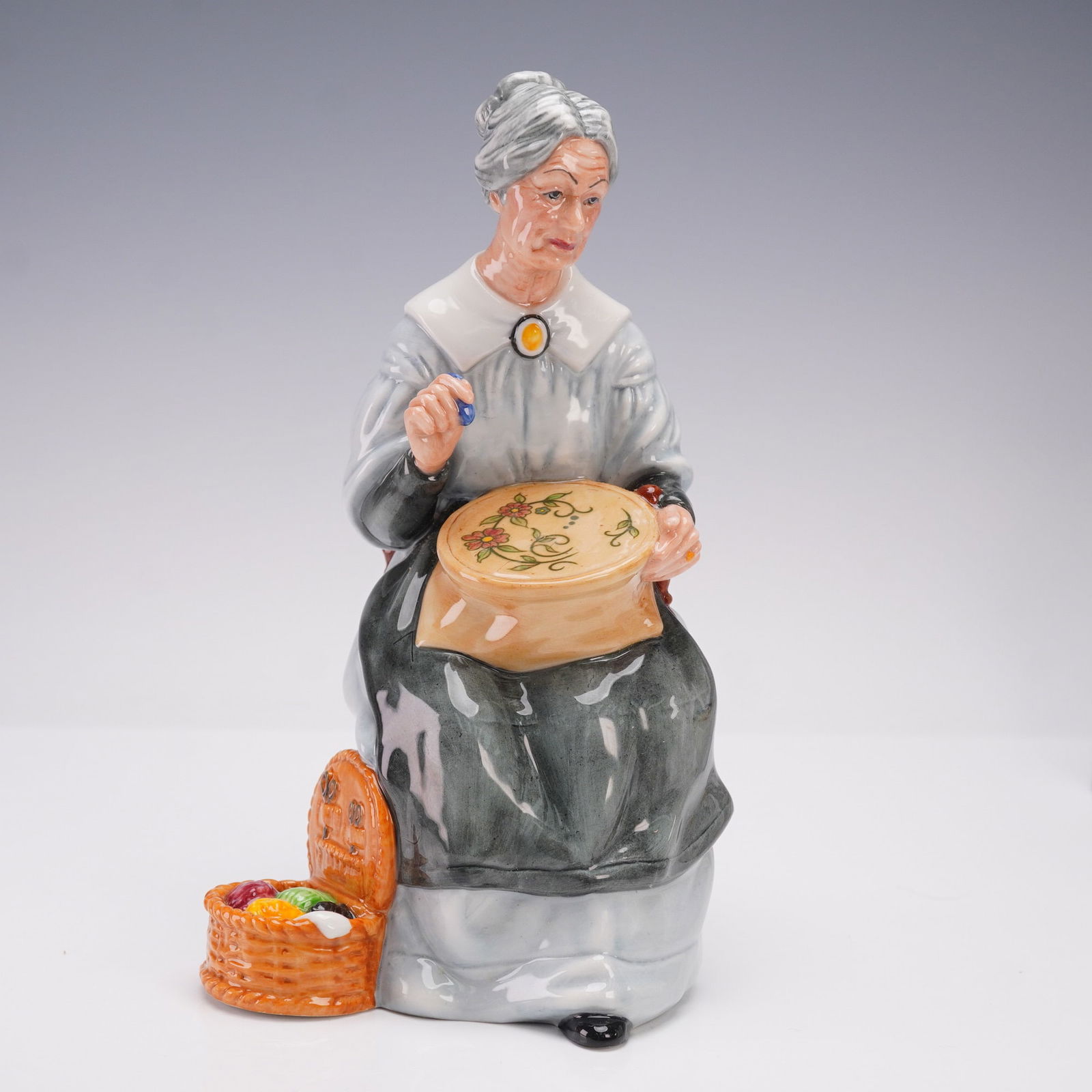 Royal Doulton Figurine, Embroidering HN2855: This porcelain hand-painted figure portrays a seated woman with grey hair neatly tied in a bun, wearing a pale blue dress with a white collar and a yellow brooch. She is shown engaged in embroidery wo