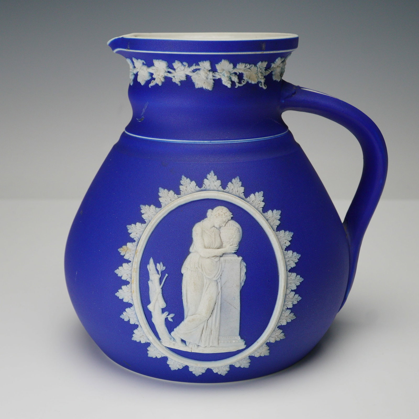 Wedgwood Blue Jasperware Pitcher: Wedgwood blue jasperware pitcher with white relief neoclassical scene in an oval cartouche and grapevine border at the neck. Rounded body with side handle and shaped spout. Wedgwood backstamp. Issued: