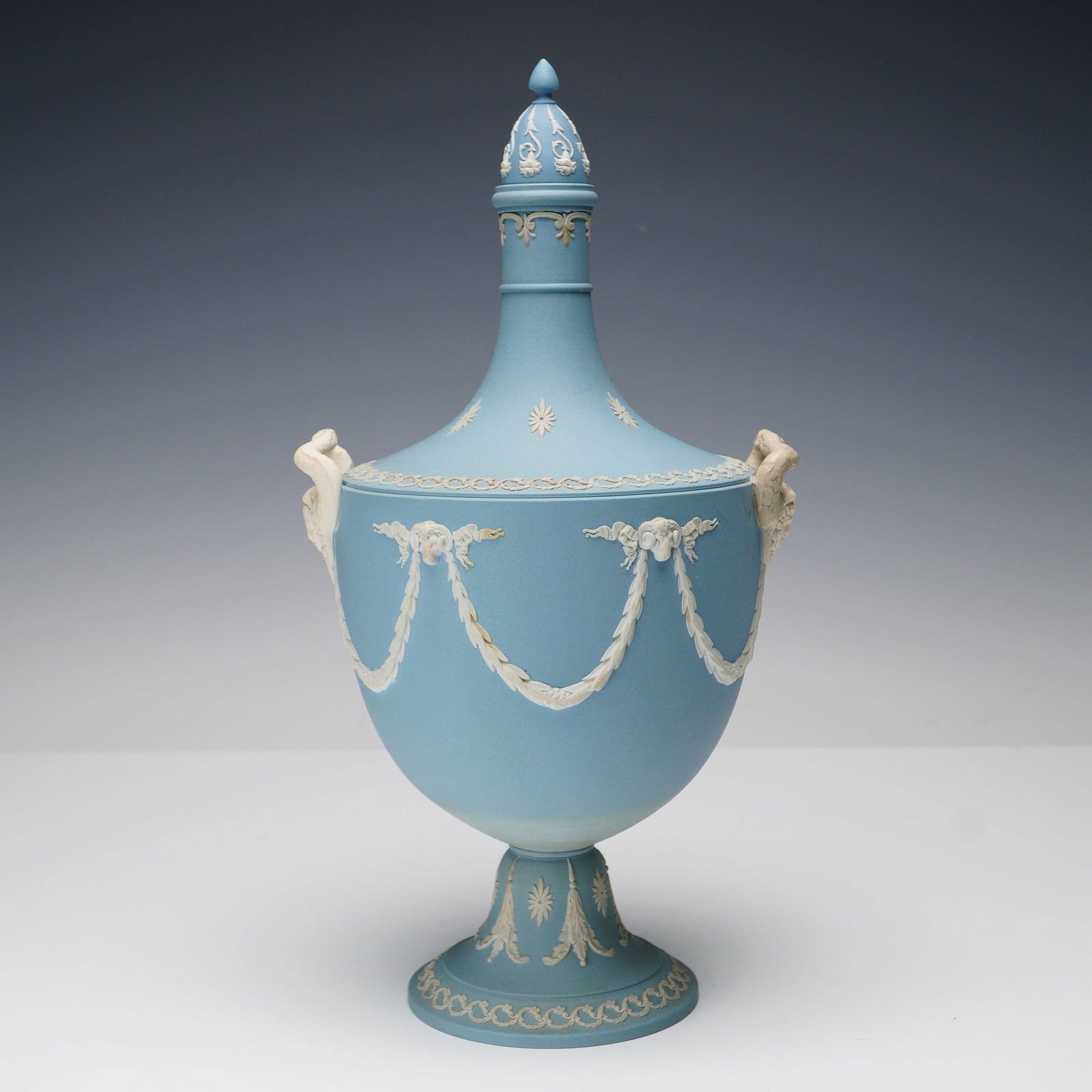 Wedgwood Blue Jasperware Lidded Urn: Wedgwood Blue Jasperware lidded urn featuring neoclassical relief decoration in white on a pale blue ground. The urn is adorned with ram's head handles joined by festooned garlands, floral motifs, and