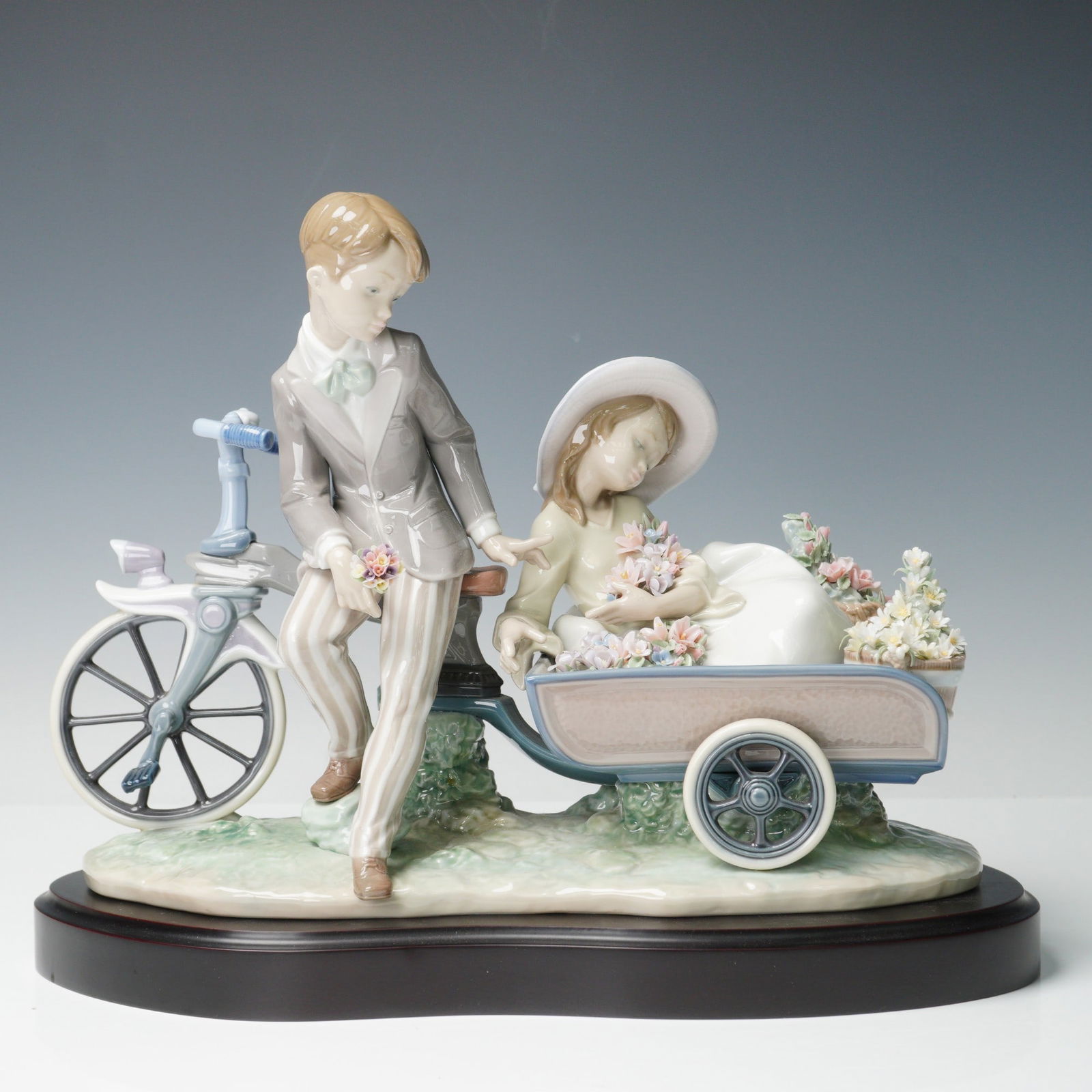 Lladro Porcelain Figurine with Base, Country Ride 1005958: Depicting a boy on a bicycle with a seated girl holding a bouquet of flowers in a cart filled with floral arrangements. The figures are dressed in pastel tones with delicate detailing, including a hat