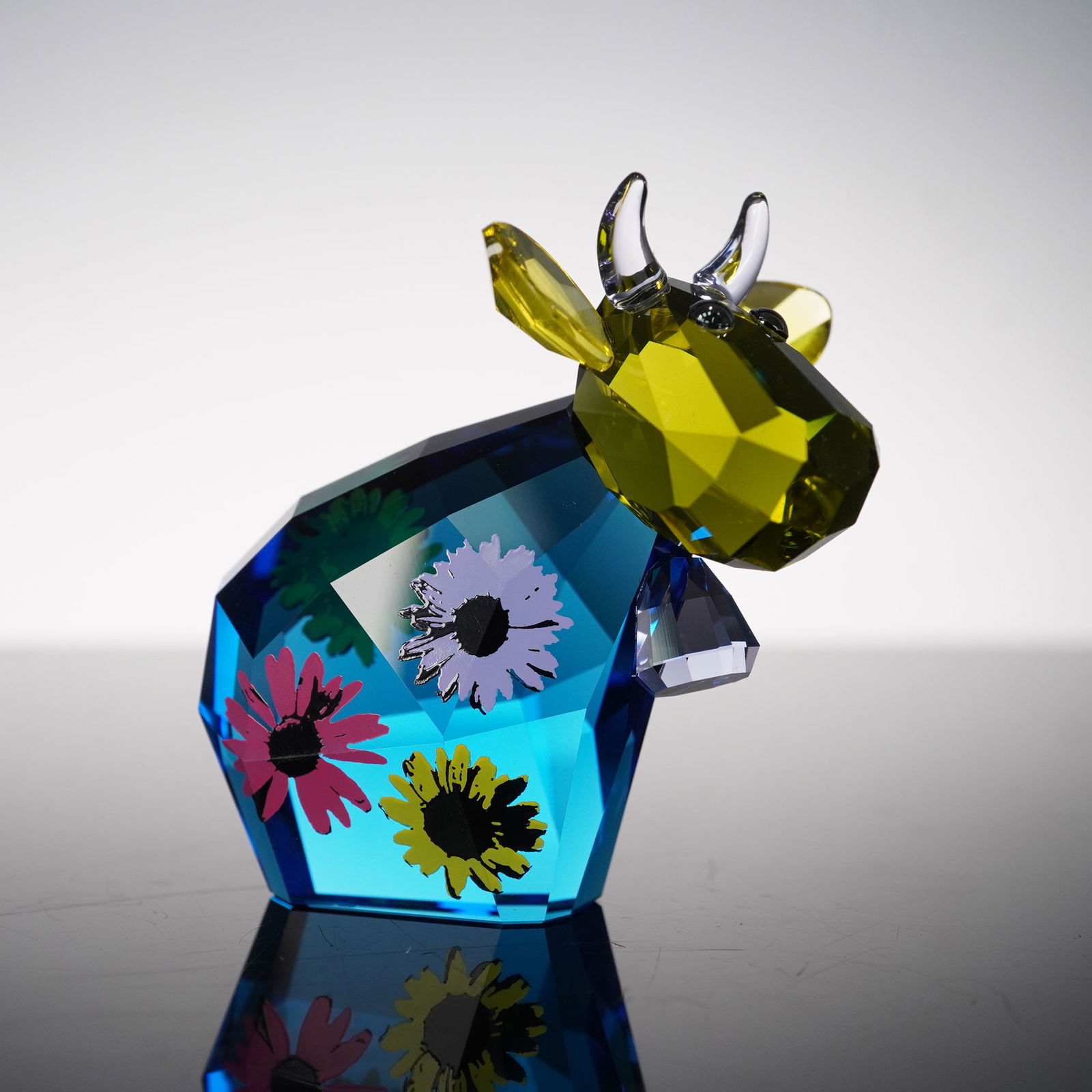 Swarovski Crystal Lovlots Figurine, Flower Power Mo: A vibrant figurine showcasing the playful cow motif adorned with faceted crystal in vivid blue tones, accented with a contrasting yellow crystal head and ears. The body features colorful printed flora