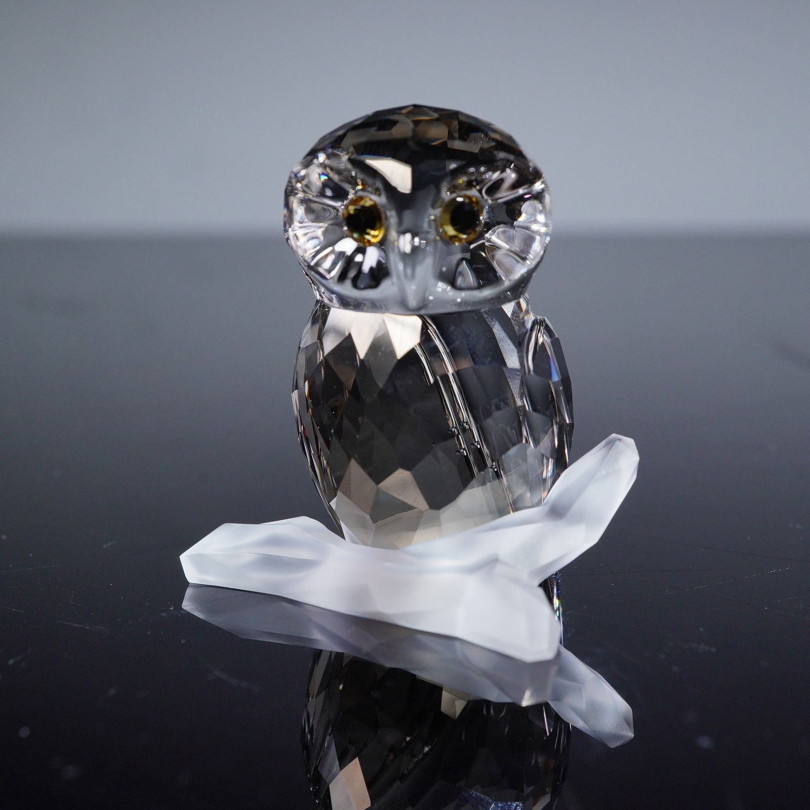 A Swarovski Crystal Owl Figurine on Frosted Branch: A Swarovski crystal owl figurine. Crafted from clear, faceted crystal, depicting a stylized owl with small, dark, possibly gold-toned eyes. The owl is perched on a separate, frosted (satin) clear crys
