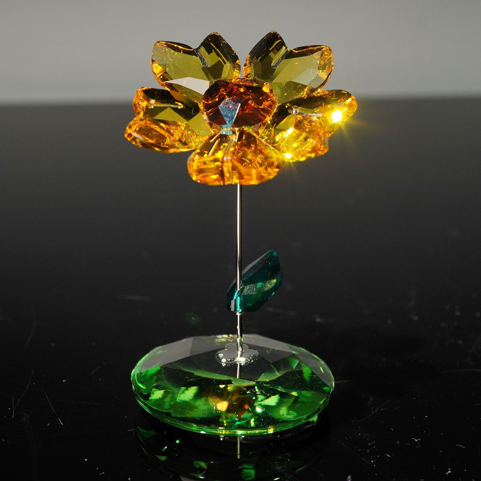 Swarovski Crystal Rocking Flower, Joy Sunflower 1016551: This crystal flower depicts a radiant sunflower rendered in golden topaz crystal with a darker crystal center. A faceted emerald satin crystal leaf adorns the silver-tone metal stem. The faceted spher