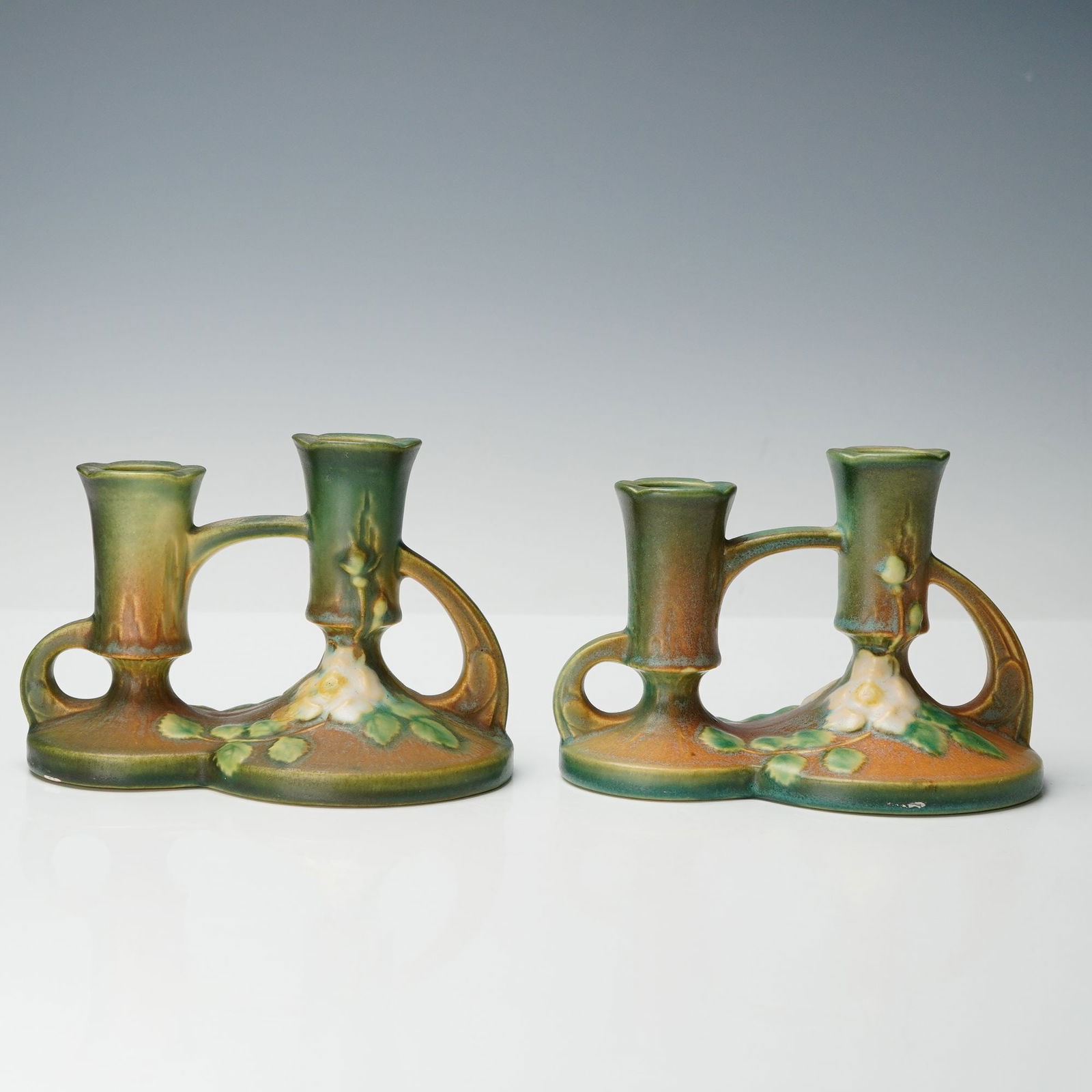 Pair of Roseville White Rose Double Candle Holders: Each piece features an intertwined dual-chamber design with a gracefully arched handle and sculpted white blossoms surrounded by green foliage on a softly textured surface of brown and moss green glaz