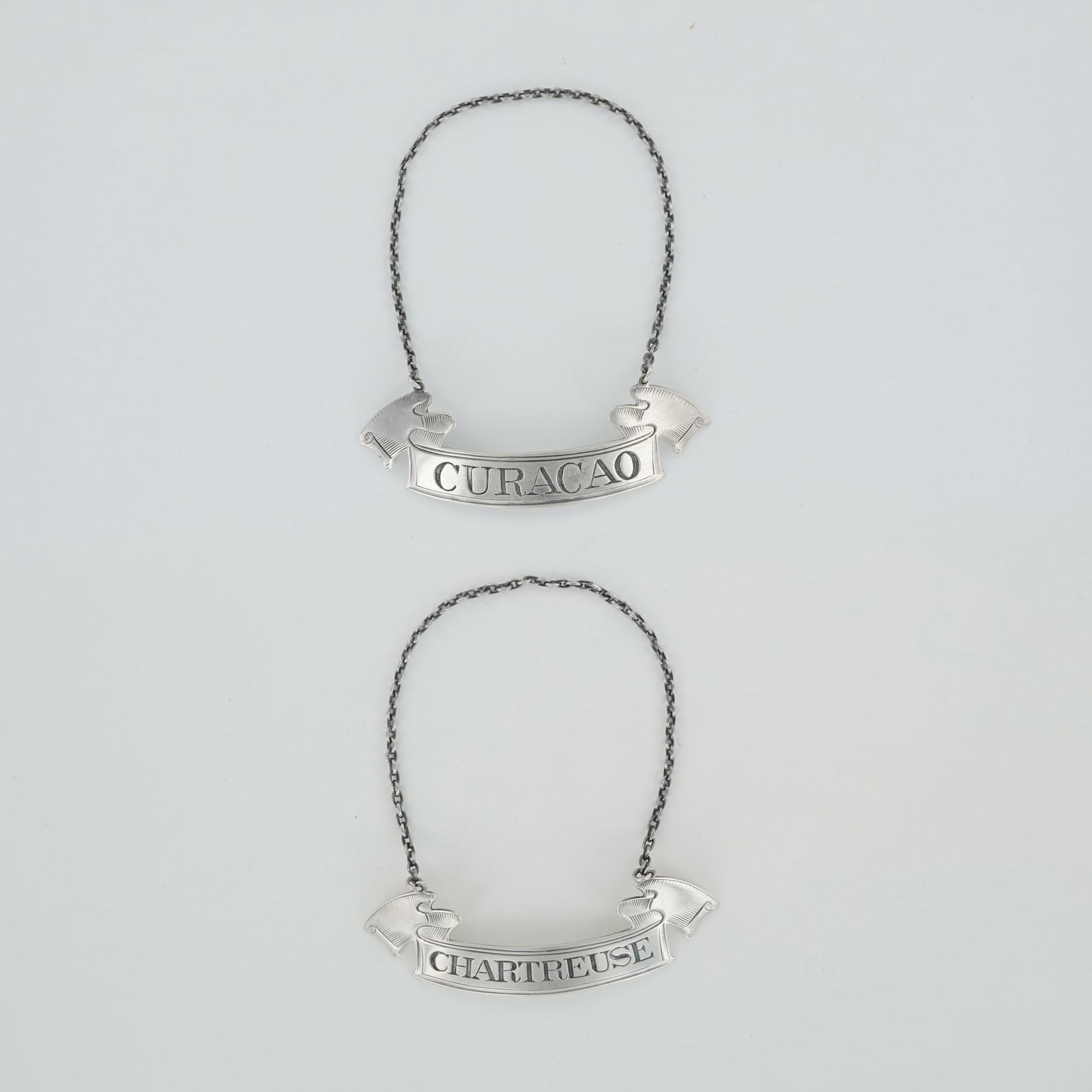 2pc Gorham Sterling Silver Decanter Labels: Each finely crafted in a banner form with flared ends and suspended from matching silver chains. One label engraved "CURACAO" and the other "CHARTREUSE," both featuring crisp lettering and decorative