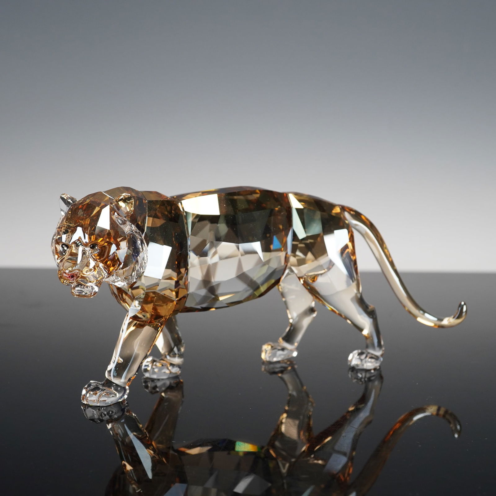 Swarovski Annual Edition, Tiger: This striking sculpture captures the majesty and strength of the tiger in a dynamic walking pose, rendered in faceted golden and clear crystal that reflects light in warm amber and honey tones. Each d
