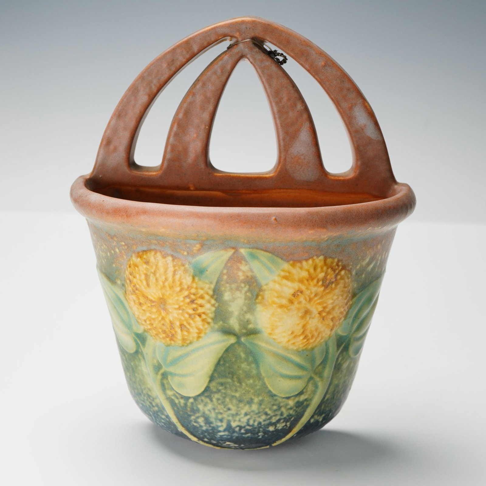 Roseville Pottery Sunflower Wall Pocket: An elegant American art pottery wall pocket featuring a rich matte glaze and relief-molded sunflower blossoms with broad green leaves against a softly shaded background transitioning from deep blue at