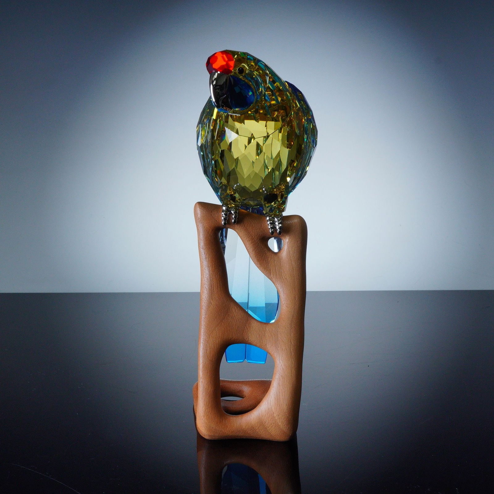 Swarovski Crystal Paradise Bird Figurine, Green Rosella: A vibrant Swarovski figurine designed by Heinz Tabertshofer. This exquisite piece features the rosella parrot in radiant green and blue crystal with red and yellow accents, perched on a stylized matte
