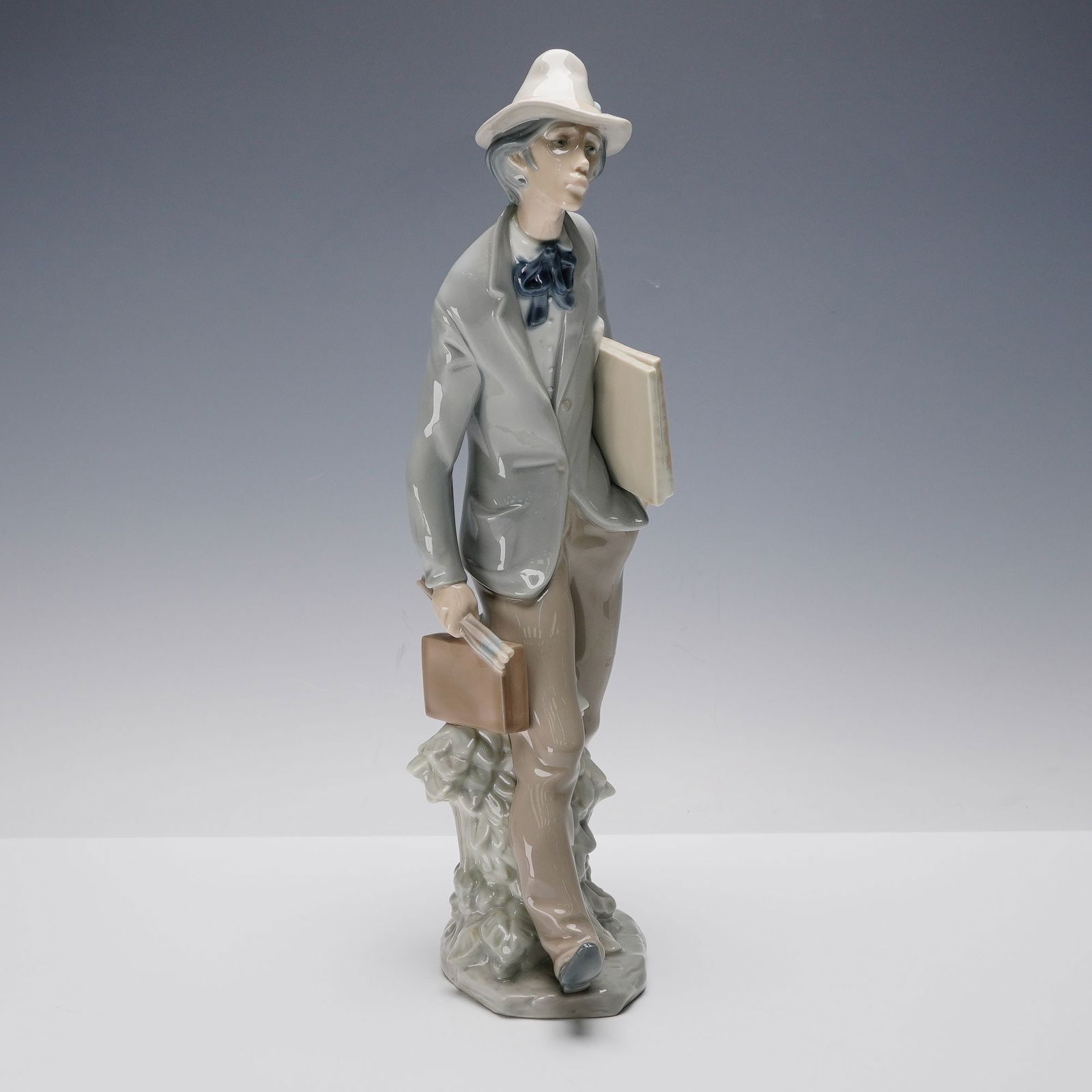 Lladro Porcelain Figurine, Artist 1004732: Glossy finish porcelain portraying a contemplative artist in hat and bow tie holding a portfolio under one arm and satchel in the other. Rendered in muted grey and beige tones with elongated stance at