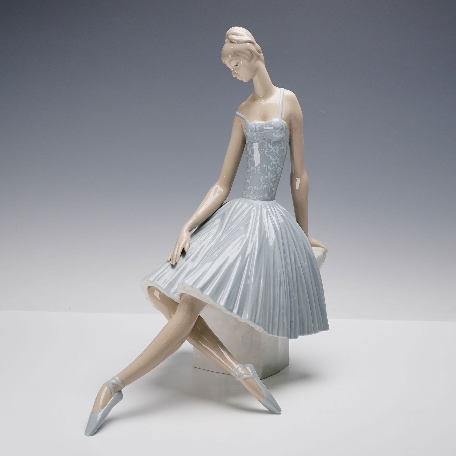 Lladro Porcelain Figurine, Ballerina Waiting Backstage 01004559: Lladro porcelain figurine titled Ballerina Waiting Backstage (01004559), designed by Fulgencio Garcia. This elegant sculpture depicts a poised young ballerina seated gracefully, one leg crossed over t