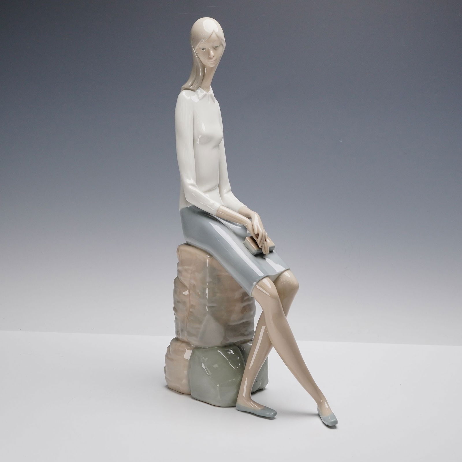 Lladro Porcelain Figurine, Girl Student 01004518: Lladro porcelain figurine titled Girl Student (01004518), designed by Fulgencio Garcia. This elegant sculpture depicts a young woman seated on stacked books, holding a small volume in her hands. Rende