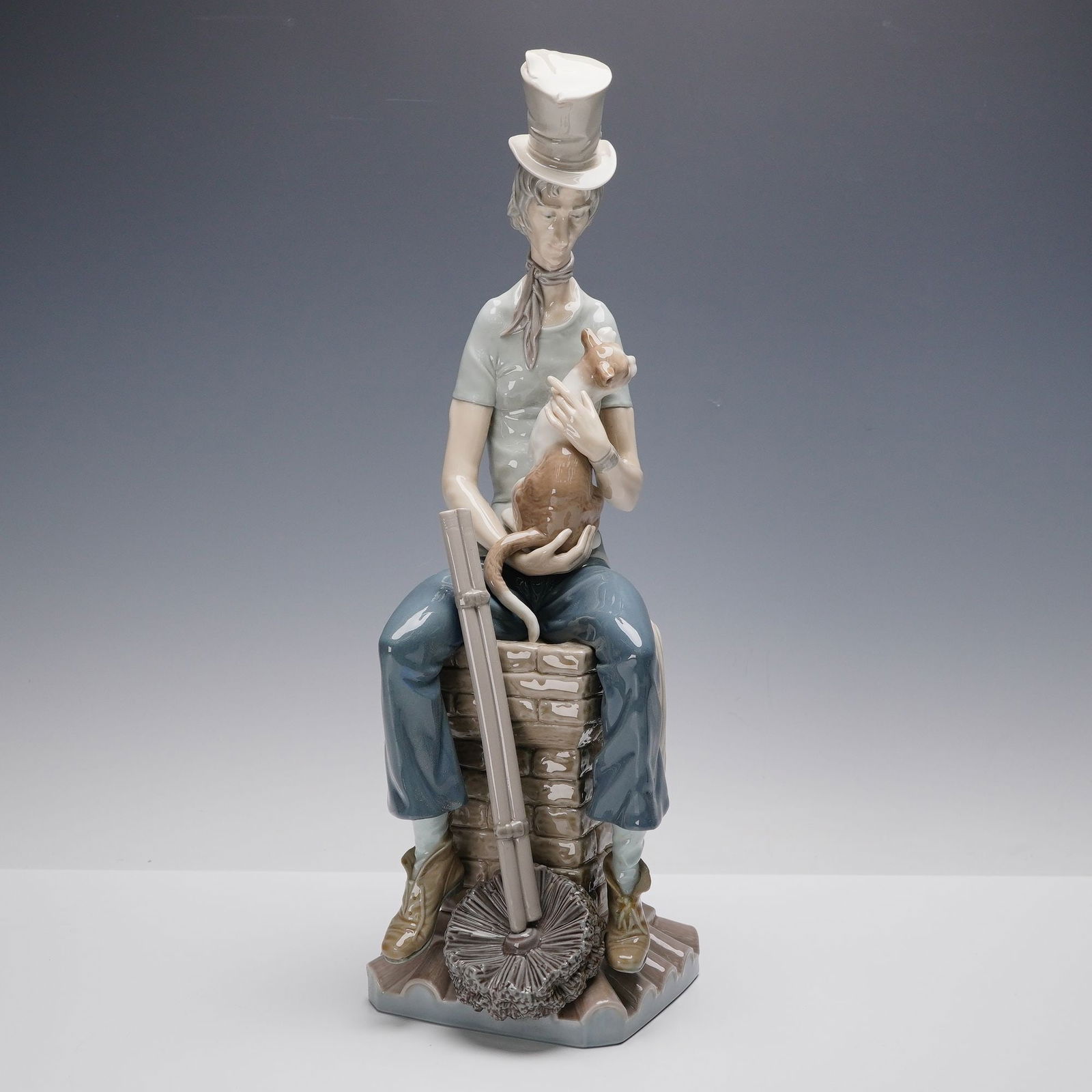 Lladro Porcelain Figurine, Sad Chimney Sweep 01001253: Lladro porcelain figurine titled Sad Chimney Sweep (01001253), designed by Salvador Furio. This charming sculpture portrays a wistful chimney sweep seated atop a brick stack, holding a small cat in hi