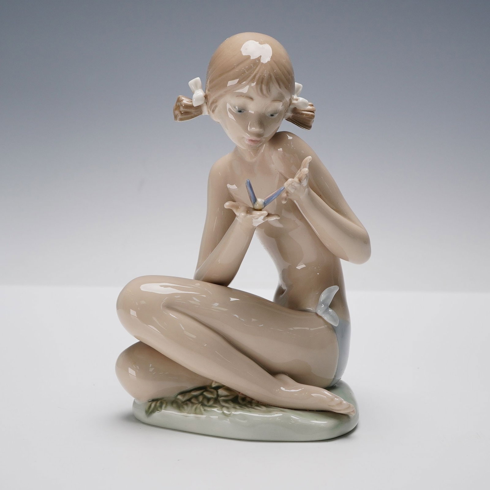 Lladro Porcelain Figurine, Free as a Butterfly 1001483: A charming porcelain figurine depicting a young girl seated cross-legged while gently holding a delicate butterfly in her hands. Her soft features and serene expression capture a moment of tender curi