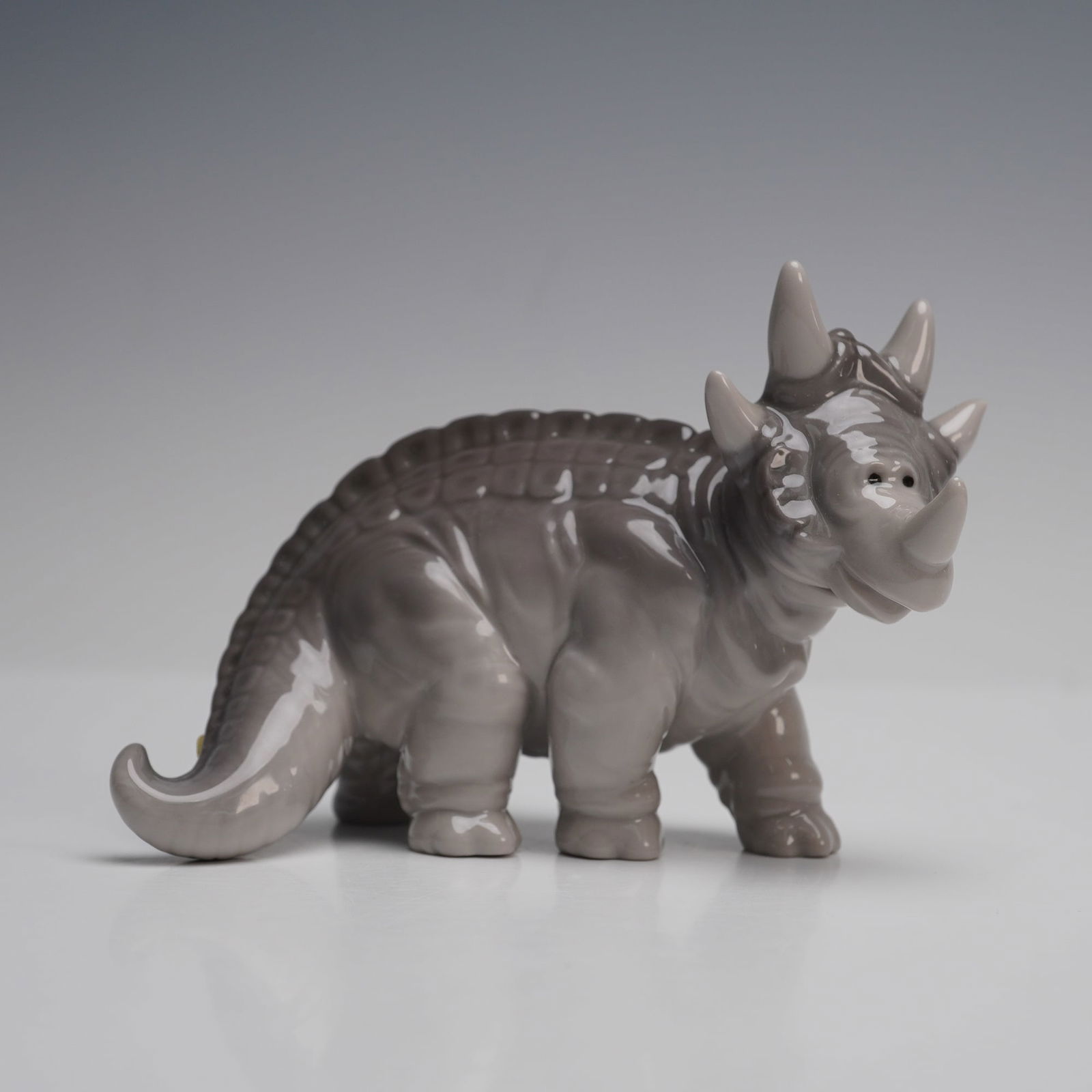 Lladro Porcelain Figurine, Spike 1007543: Depicting a triceratops dinosaur, crafted in a smooth glazed gray finish. The piece features detailed sculpting that emphasizes the animal's characteristic horns, frill, and textured body. The undersi