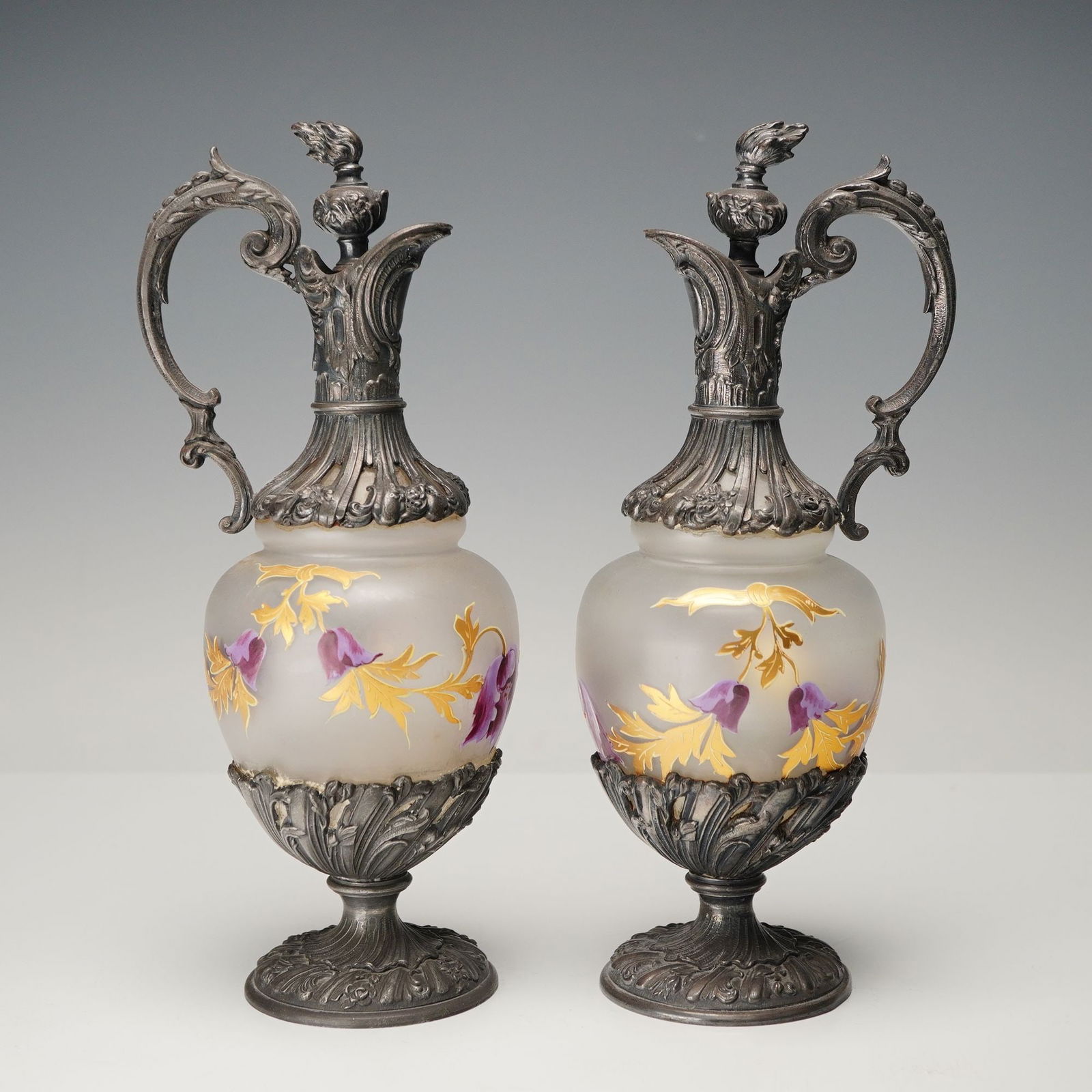 Pair of Hand Painted Frosted Glass and Metal Ewers with Stoppers: Elegant ewers with ornate cast metal mounts and handles, each hand-painted with vivid purple and gold floral motifs. The frosted bodies feature delicate gilt scrollwork and applied detailing, compleme