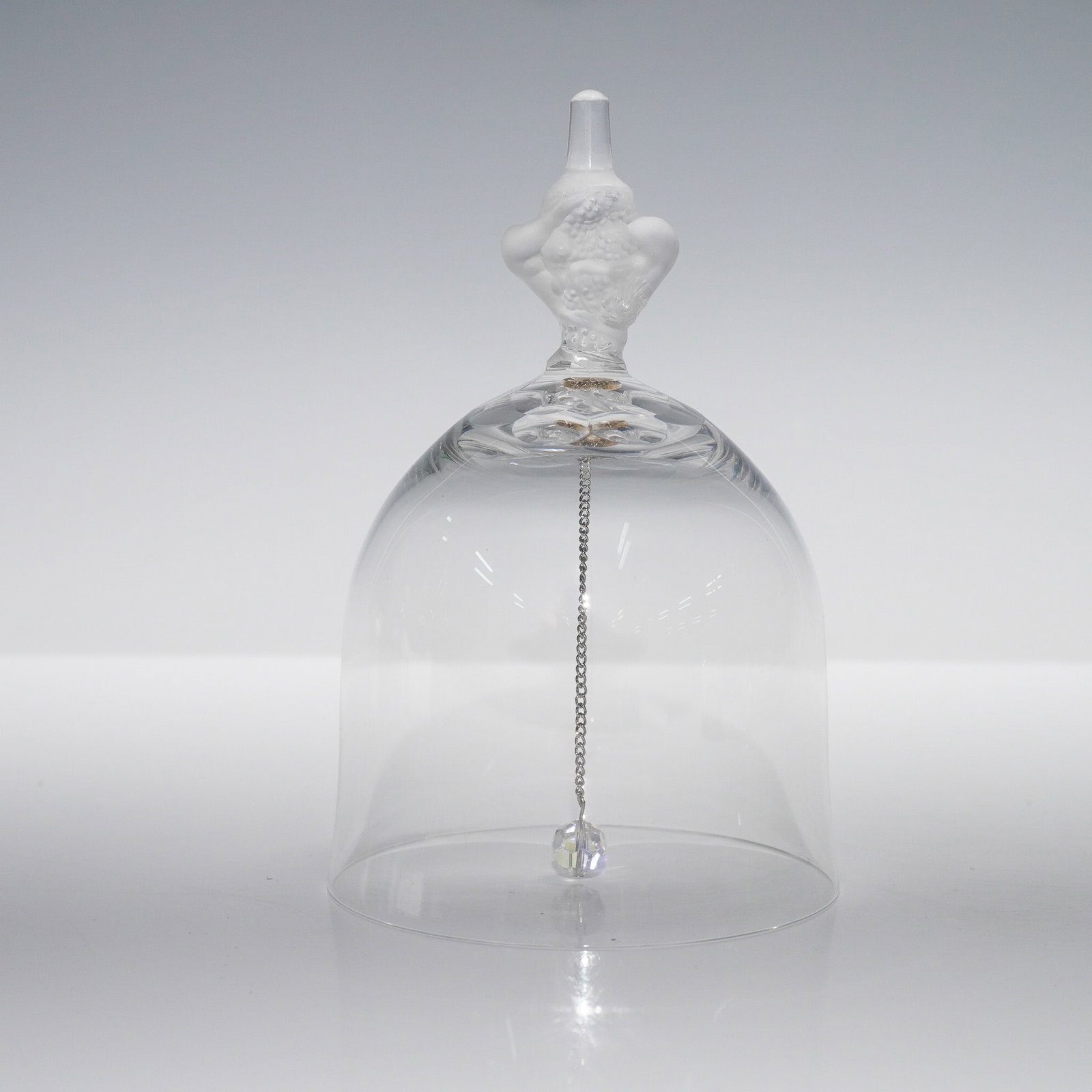 Crystal Bell with Frosted Handle: Clear crystal bell featuring a domed body with a suspended faceted clapper on a silver-tone chain. The handle is finely sculpted in frosted glass, depicting a pair of cherubs surrounded by fruit, in a