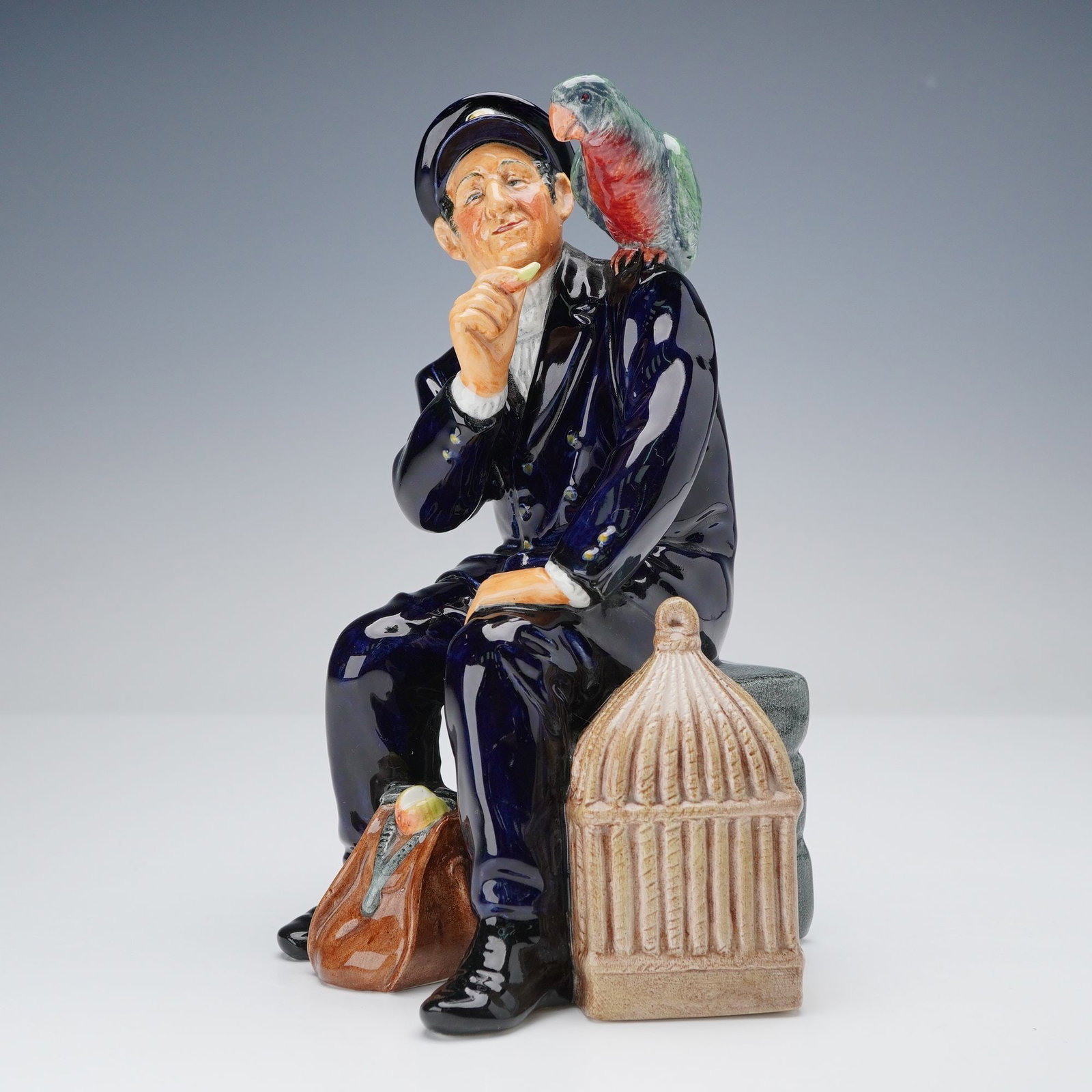 Royal Doulton Figurine, Shore Leave HN2254: An hand-painted porcelain figurine of a cheerful sailor seated on a stone dock with a colorful parrot perched on his shoulder, a birdcage by his side, and a brown duffel bag resting at his feet. The f