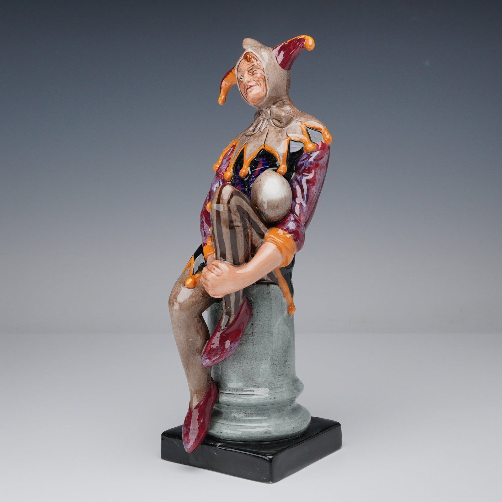 Royal Doulton Figurine, The Jester HN2016: The Royal Doulton figurine The Jester HN2016 depicts a lively jester in vibrant red, orange, purple, and gold attire, seated on a stone column. Modeled by Charles Noke, the figurine showcases expressi