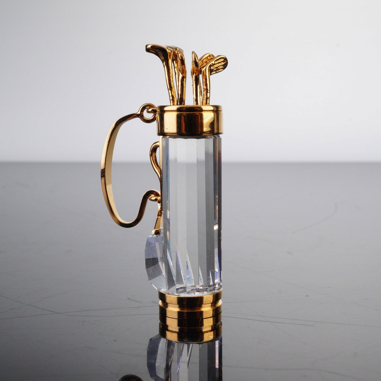 Swarovski Crystal Memories Figurine, Golf Bag: The figure is formed as a golf bag from clear faceted crystal fitted with gold tone metal clubs, handle, and end caps. The construction shows the vertical faceted body, attached metal carry handle, an