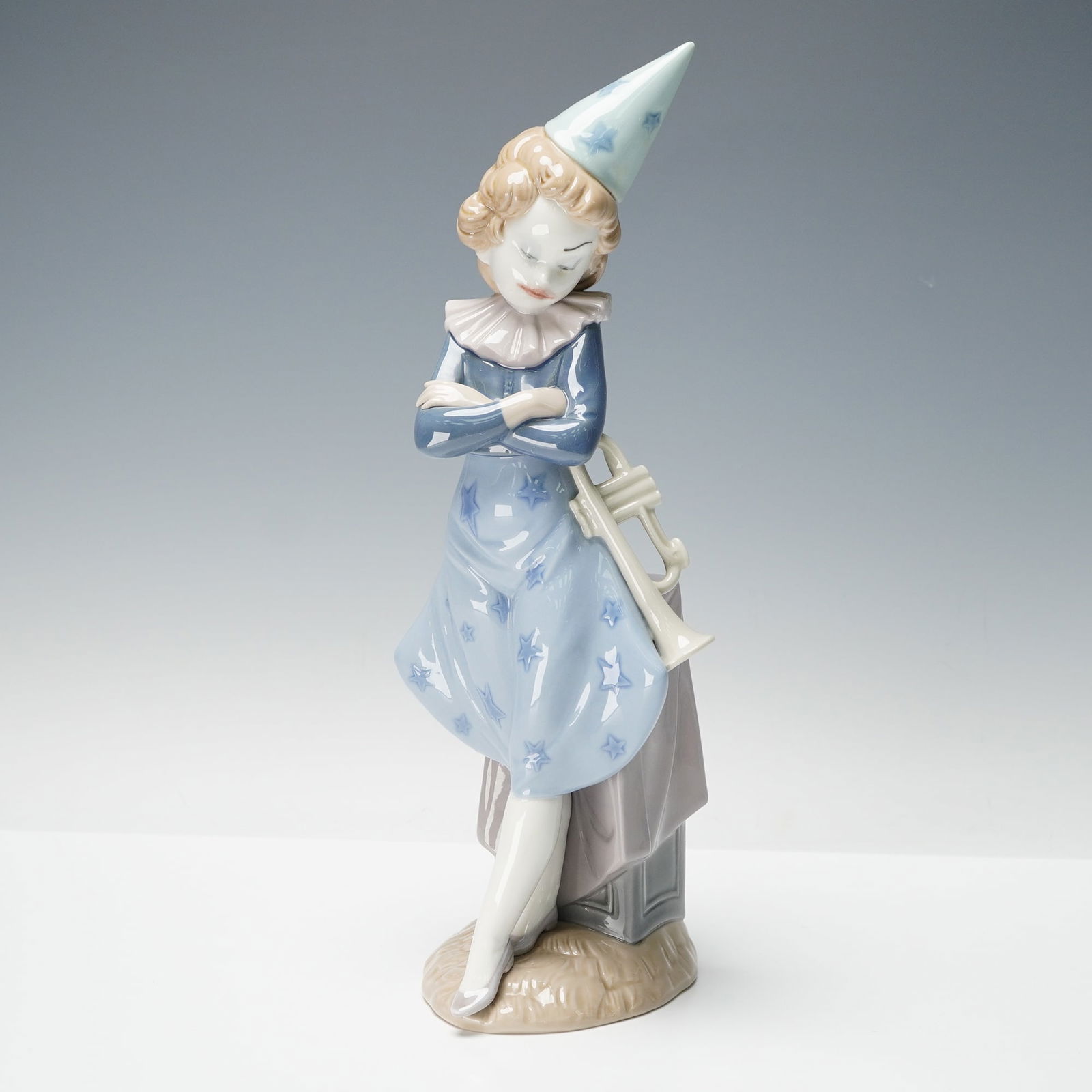 Lladro Porcelain Clown with Trumpet Sculpture 01005060: Designed by Salvador Debon the piece depicts a young clown figure in a star-patterned blue outfit, standing with arms crossed and holding a trumpet. Finished in Lladro's signature high-gloss glaze. Ma