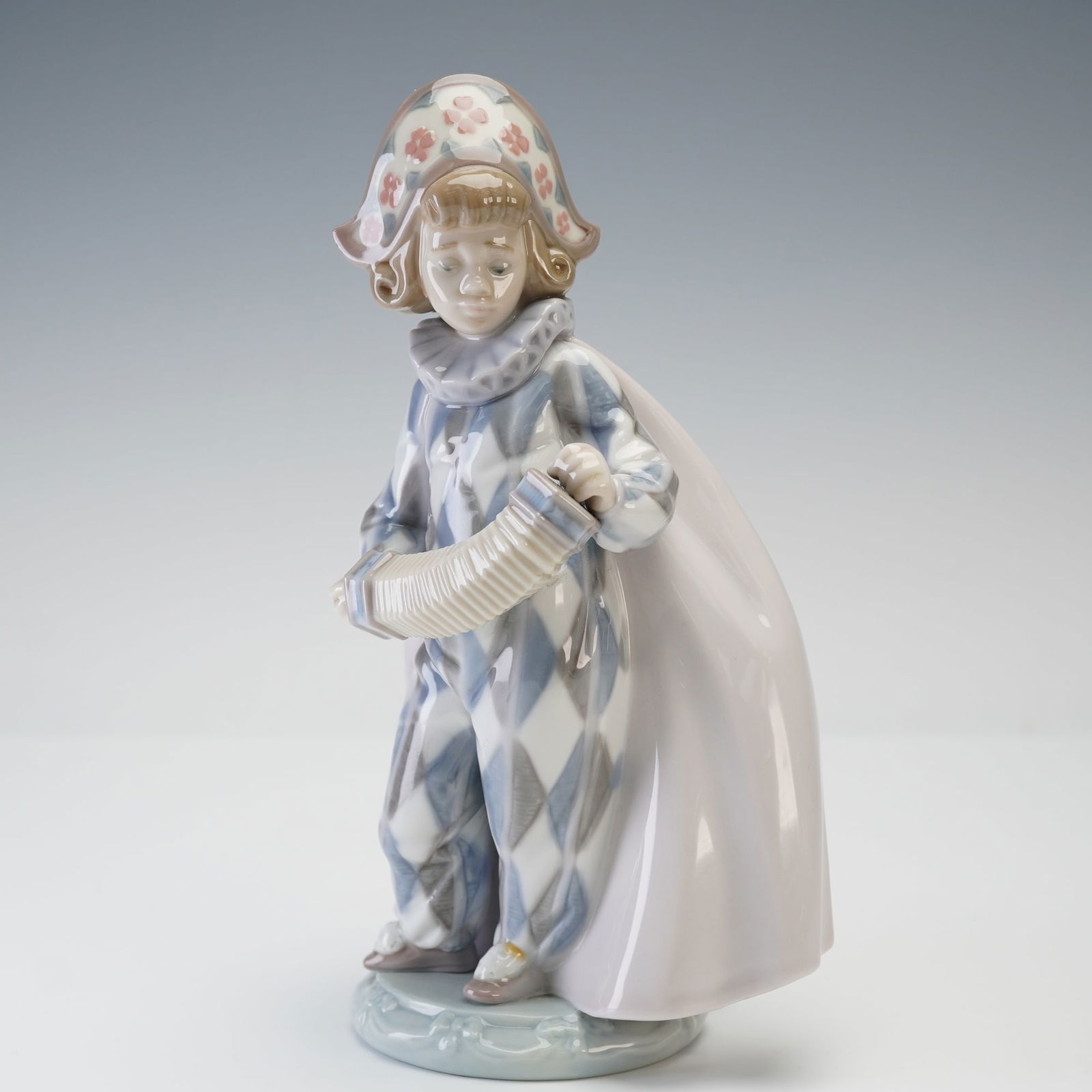 Lladro Porcelain Figurine, Concertina 1005695: Charming Lladro porcelain figurine titled Concertina, model 1005695, depicting a young harlequin in a pastel blue and white diamond-patterned costume, holding a concertina accordion. The figure is fin
